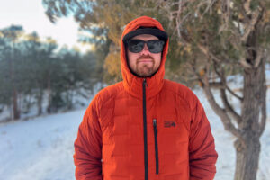 A person wearing sunglasses and the Mountain Hardwear StretchDown Hoody with the hood up, standing in a snowy forest.