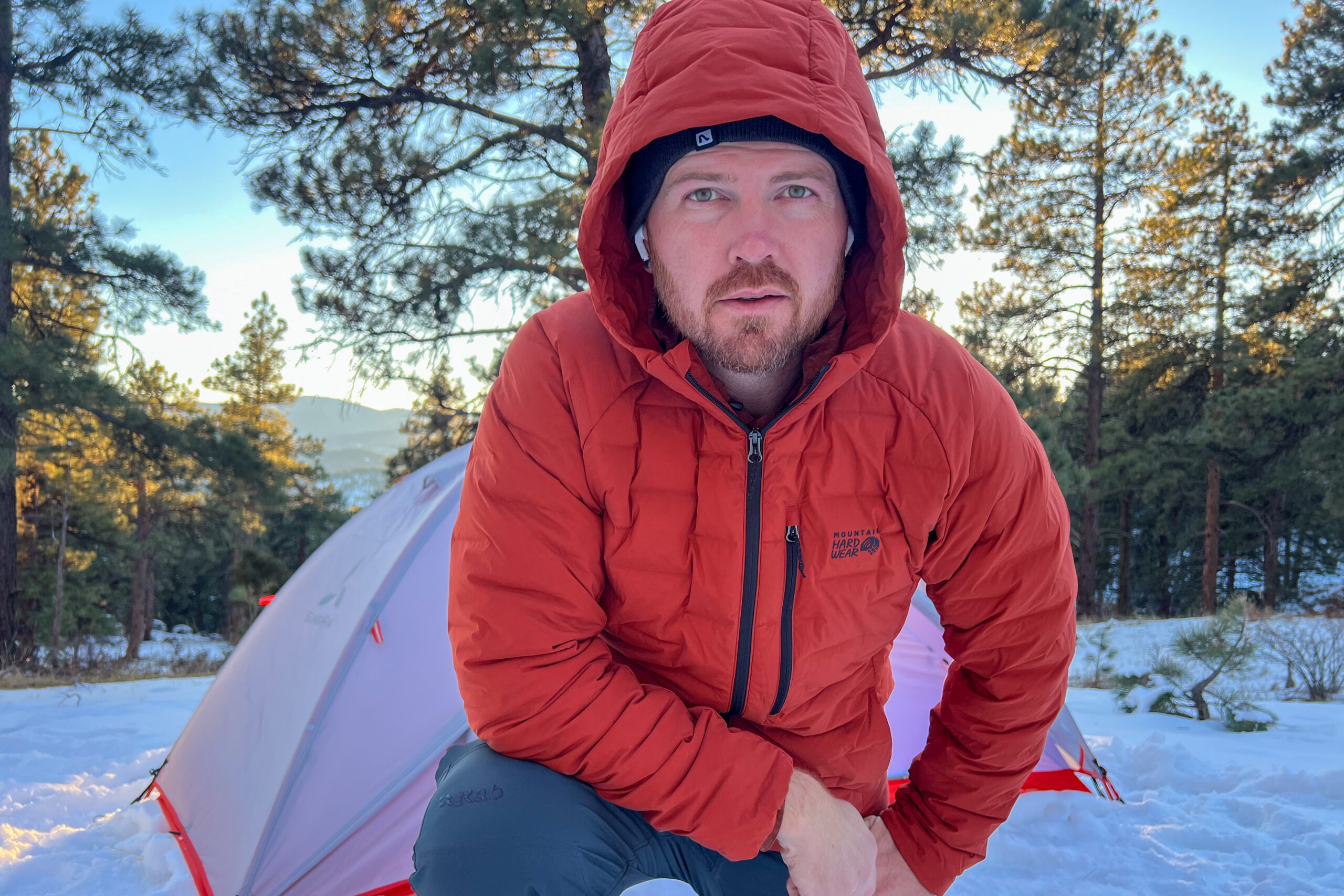 A person wearing the Mountain Hardwear StretchDown Hoody kneeling in front of a tent, with snow and pine trees in the background.