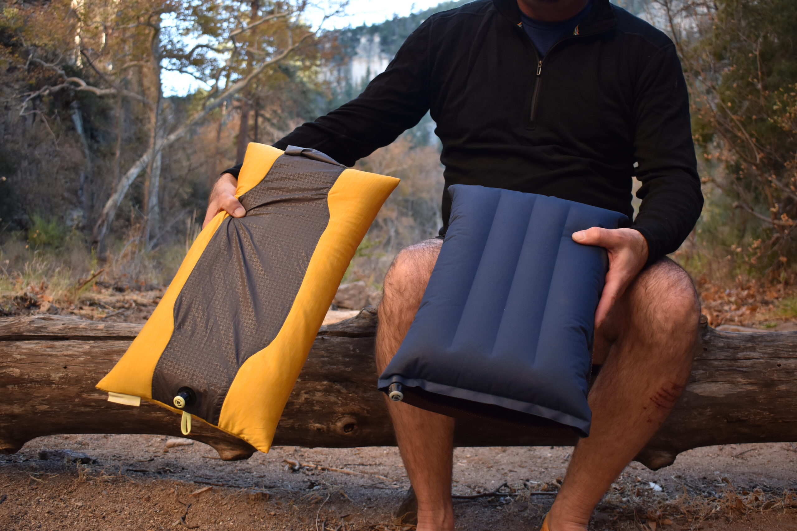 A backpacker sitting on a log holding the thinner and wider NEMO Fillo Elite Luxury pillow in his right hand and smaller, thicker NEMO Fillo pillow in his left hand