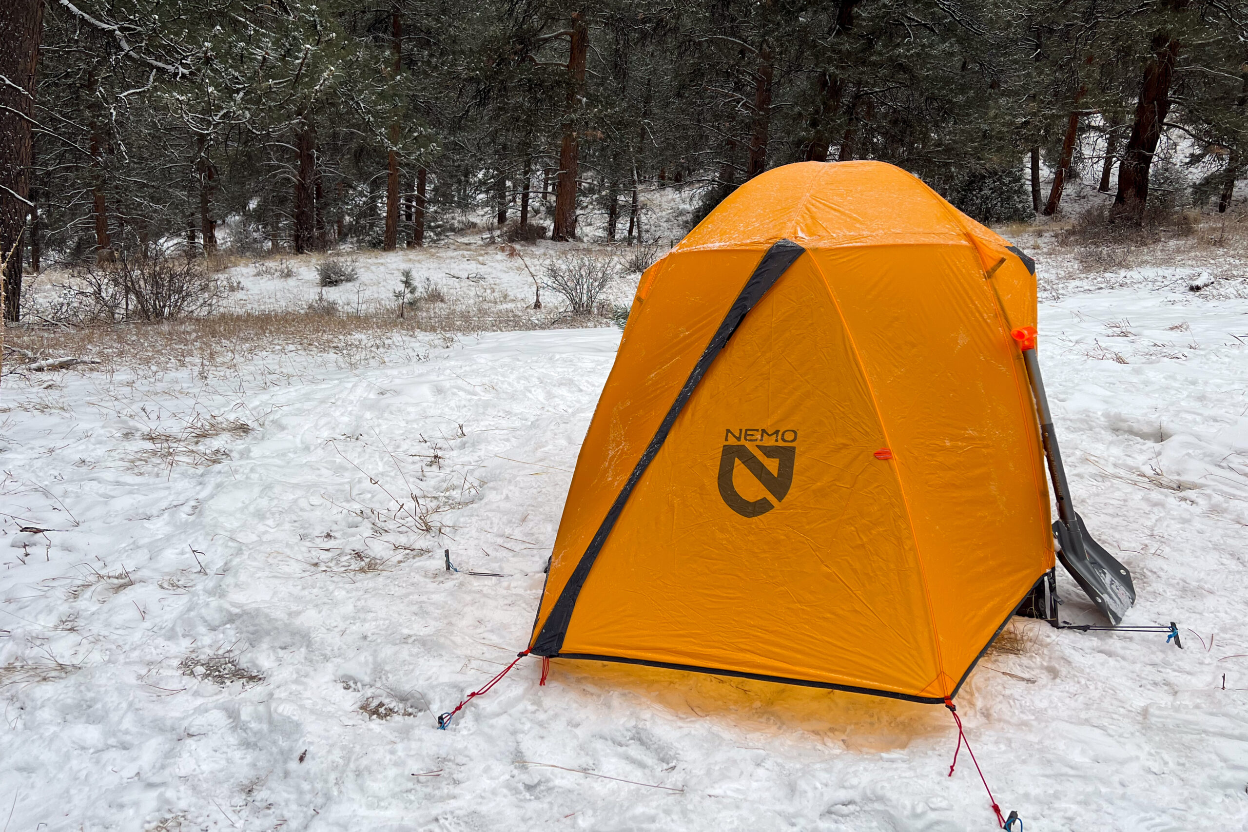 A Nemo Kunai 2P tent fully covered with its orange rainfly, set up in a snowy clearing with a snow shovel propped against it and trees in the background.