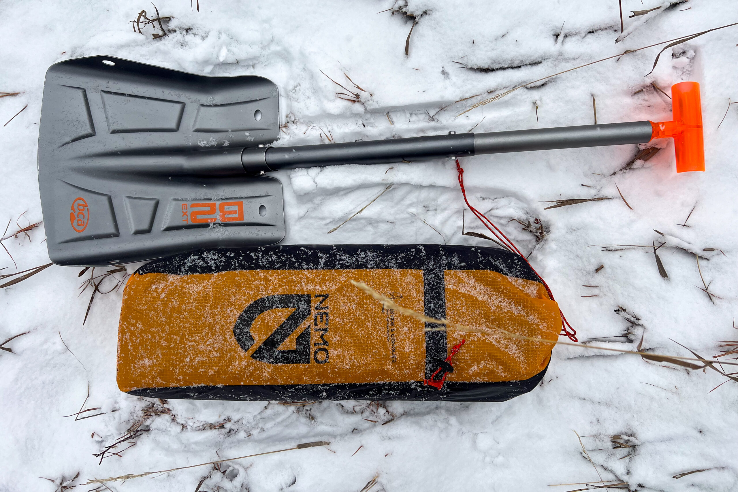 The Nemo Kunai 2 tent packed in its orange storage bag alongside a gray avalanche shovel, both lying on snow-covered ground.