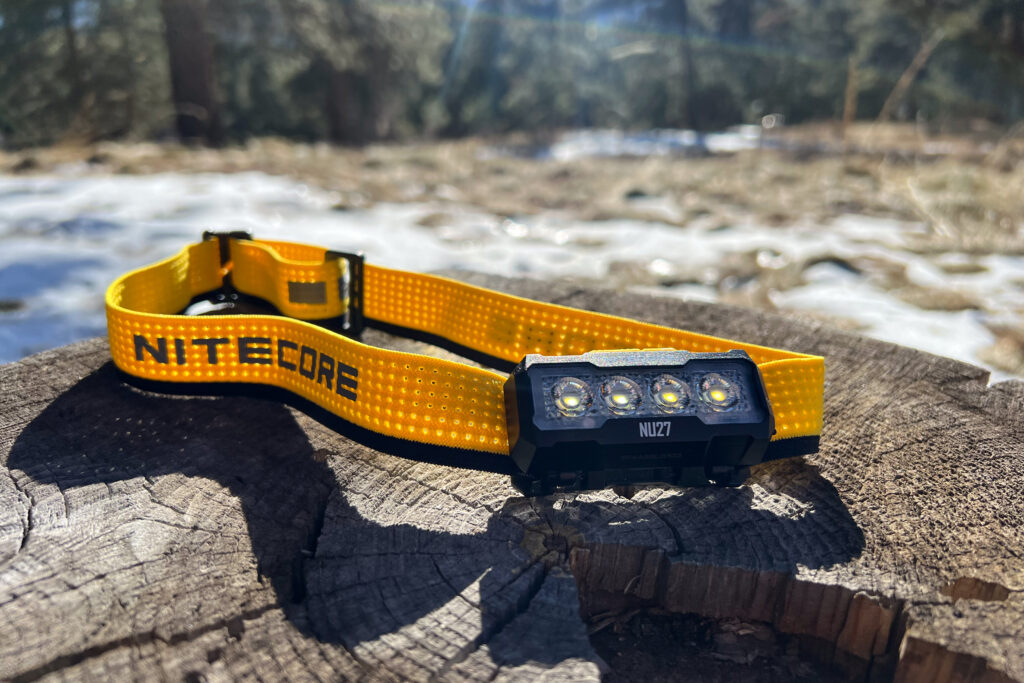 The photo features the Nitecore NU27 headlamp sitting on a log in a forest setting.