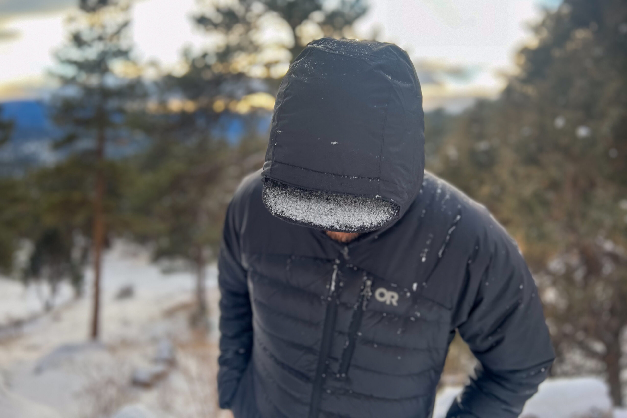 A person standing in the snow, wearing the Outdoor Research Helium Down Hoody zipped up with the hood on, looking straight at the camera.