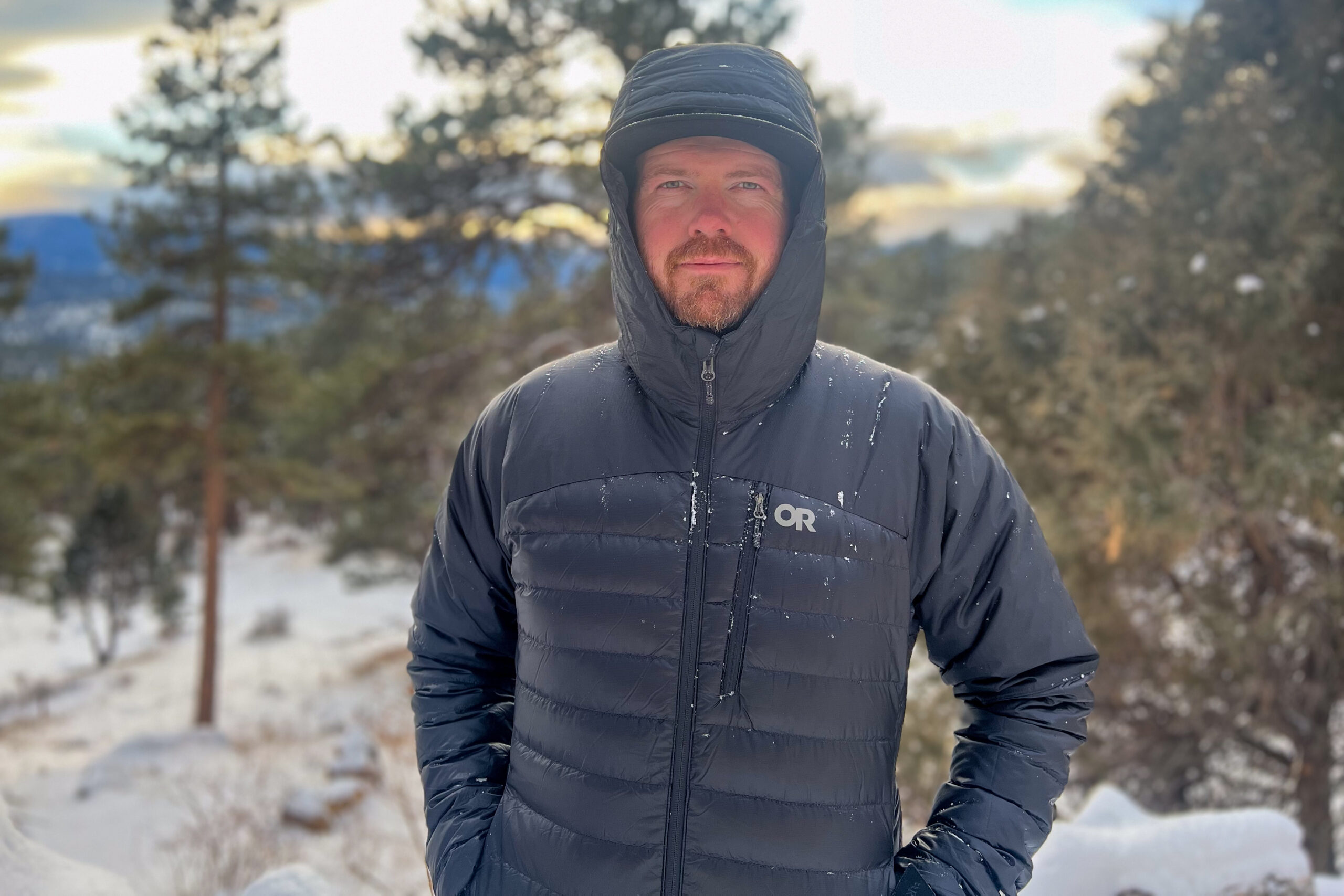 A person wearing the Outdoor Research Helium Down Hoody with snow falling on the hood and shoulders, set in a serene snowy forest.