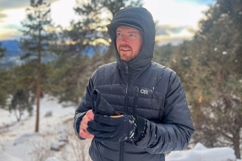 A person wearing the Outdoor Research Helium Down Hoody with the hood up, surrounded by a snowy forest landscape.