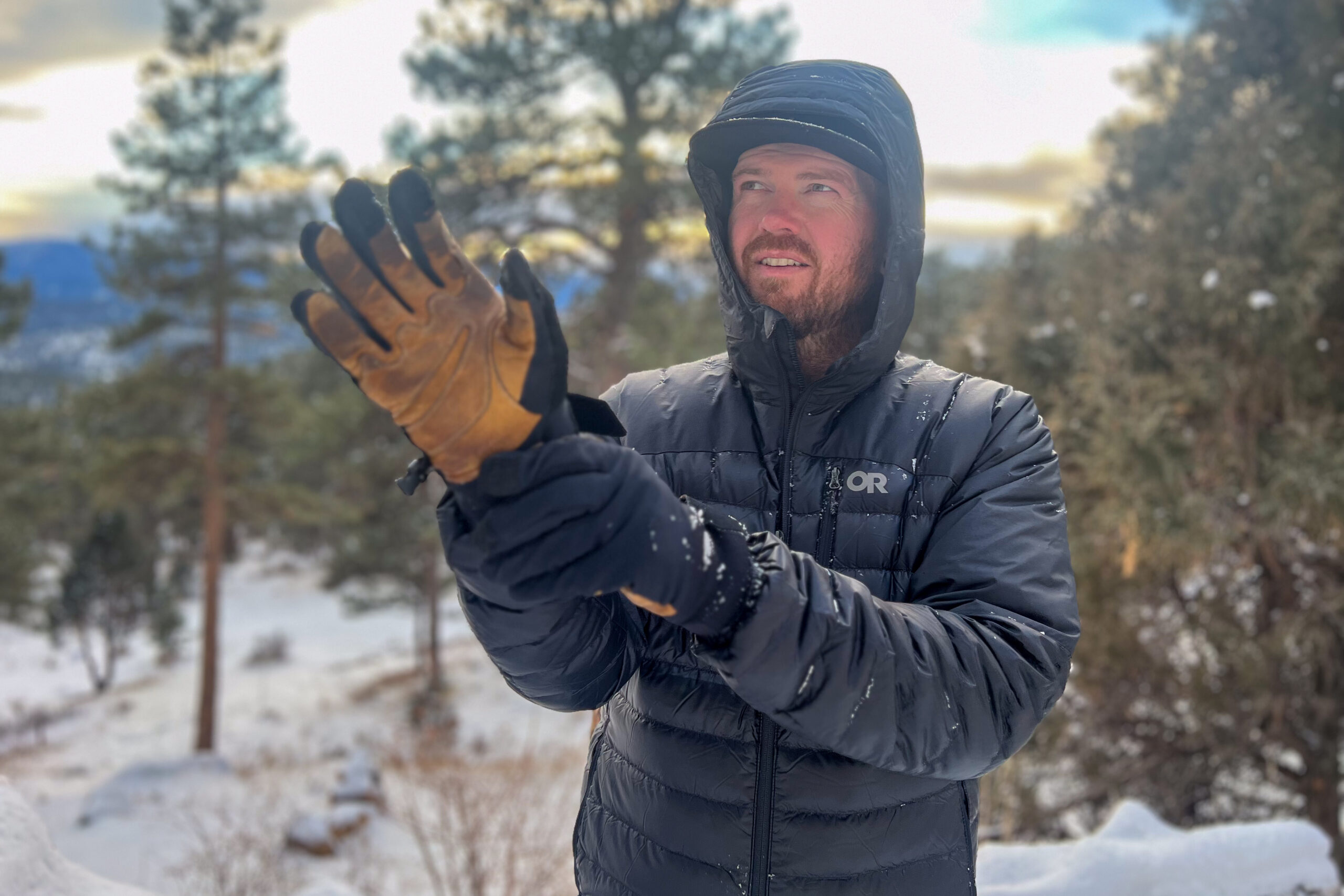 A person wearing the Outdoor Research Helium Down Hoody while putting on gloves, with snow visible on the jacket in a winter forest setting.