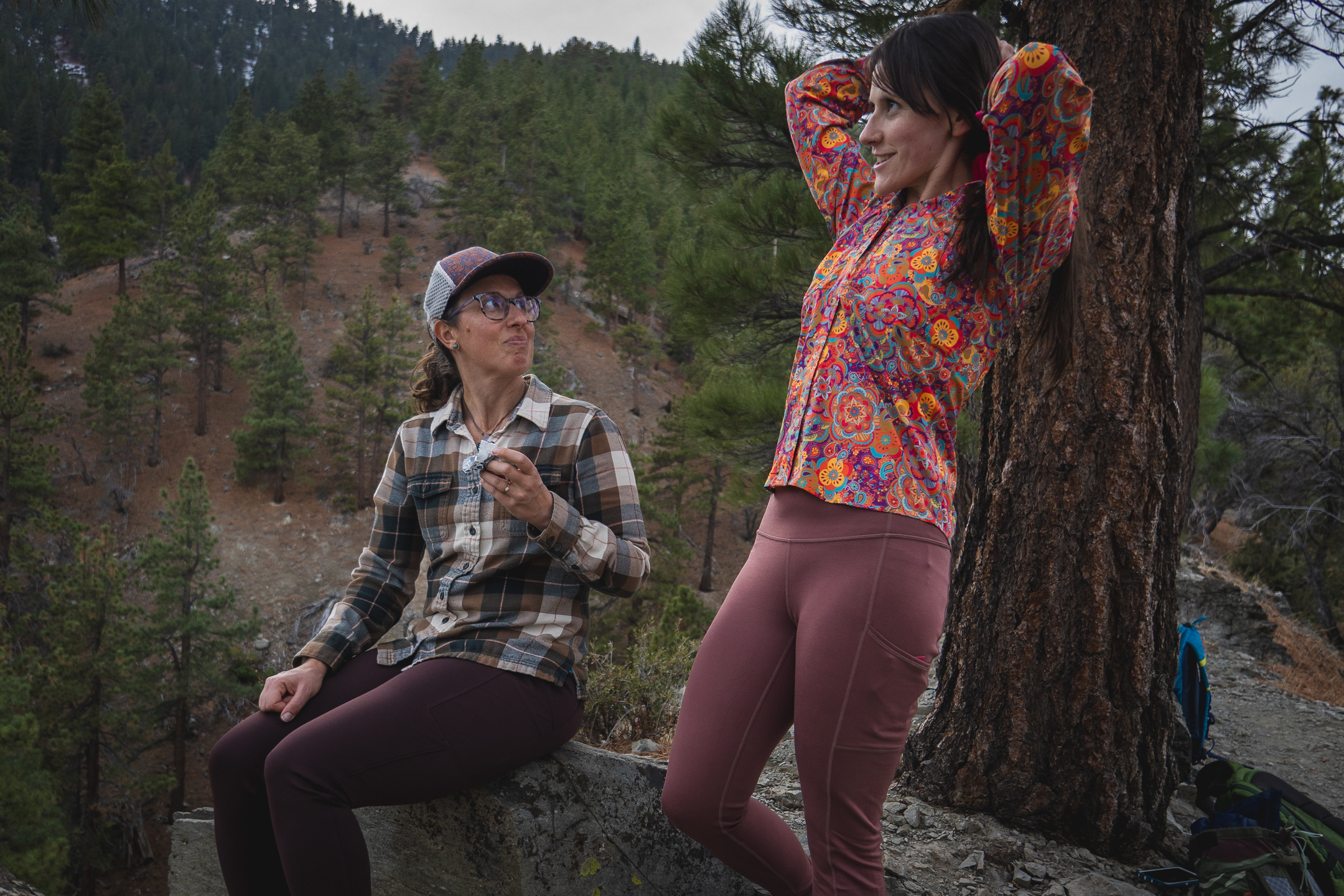 two women talking while on a hike. one is sitting in dark brown pants and a matching flannel top, wearing a hat and glasses, and the other is standing, wearing pastel pink leggings and a bright multicolored top