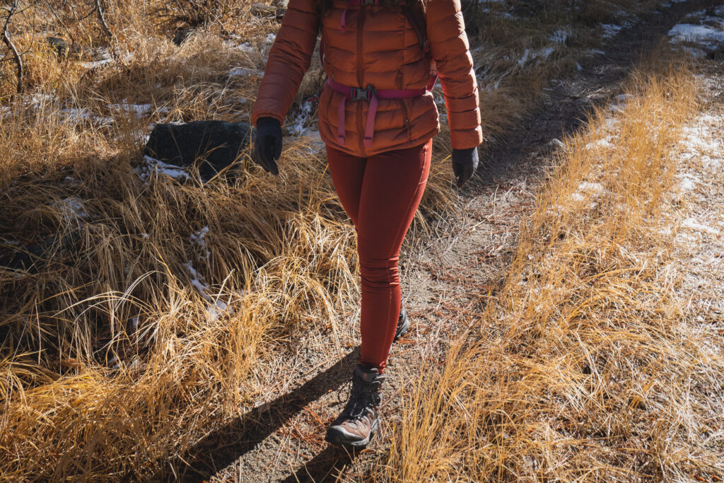 Person in reddish/orange leggings, with dry grass in background, wearing a bronze colored down jacket and black gloves