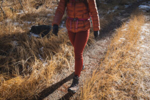 Person in reddish/orange leggings, with dry grass in background, wearing a bronze colored down jacket and black gloves