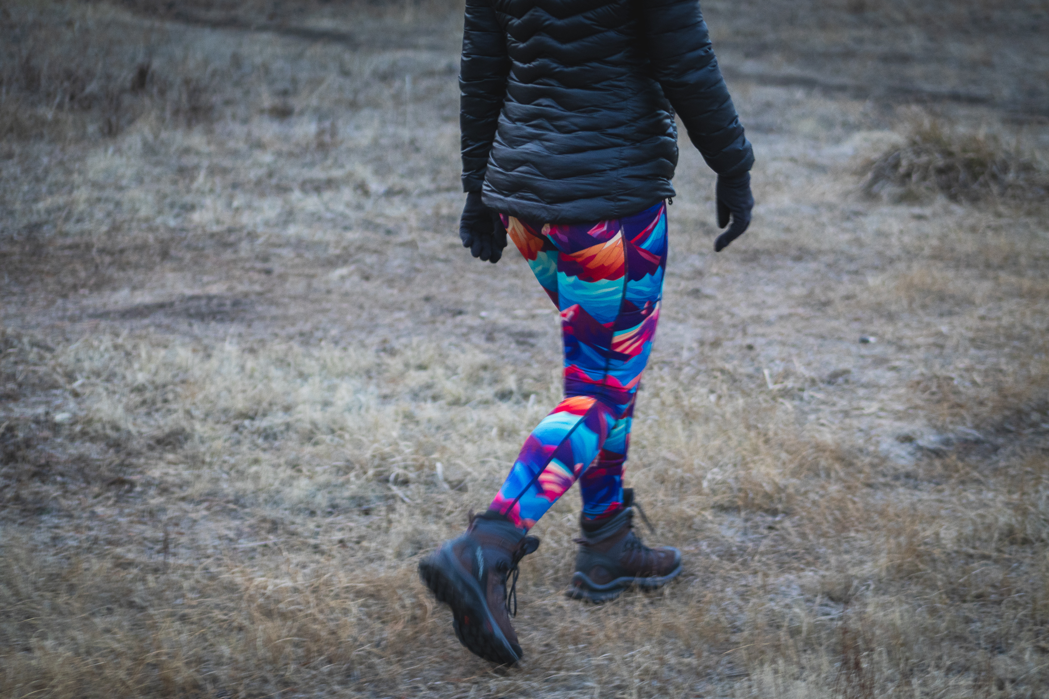 Woman in bright colored leggings (blue, purpled, orange) that have patterns of mountains, hiking on a trail in a meadow in dry grass. Person is wearing a black puffy jacket, black thin gloves and brown and grey hiking boots.