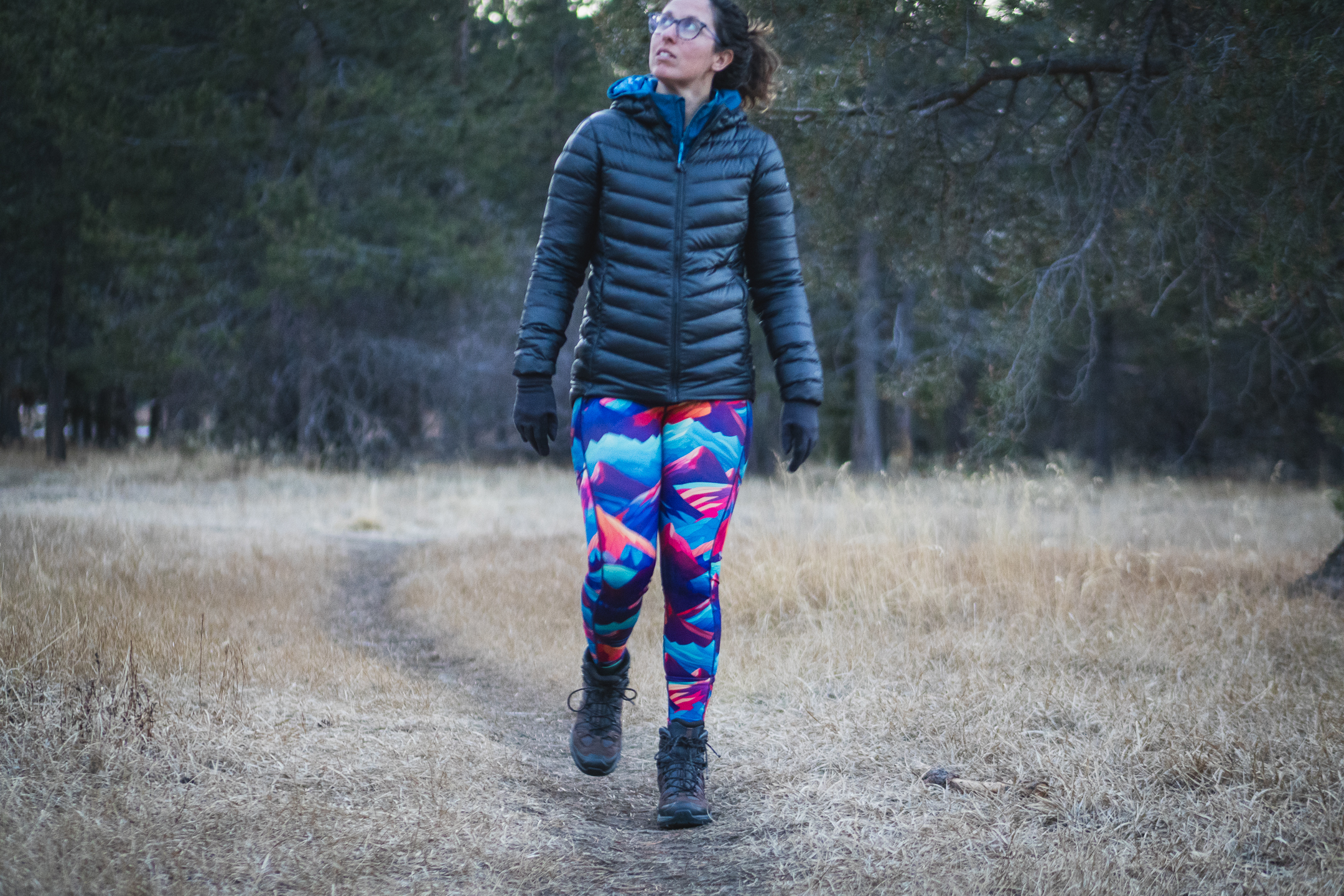 Woman in bright colored leggings (blue, purpled, orange) that have patterns of mountains, hiking on a trail in a meadow in dry grass. There is a thin windy trail going into the background, and trees blurred in the background. Person is looking up and out of the frame, and is wearing a black puffy jacket and brown and grey hiking boots.