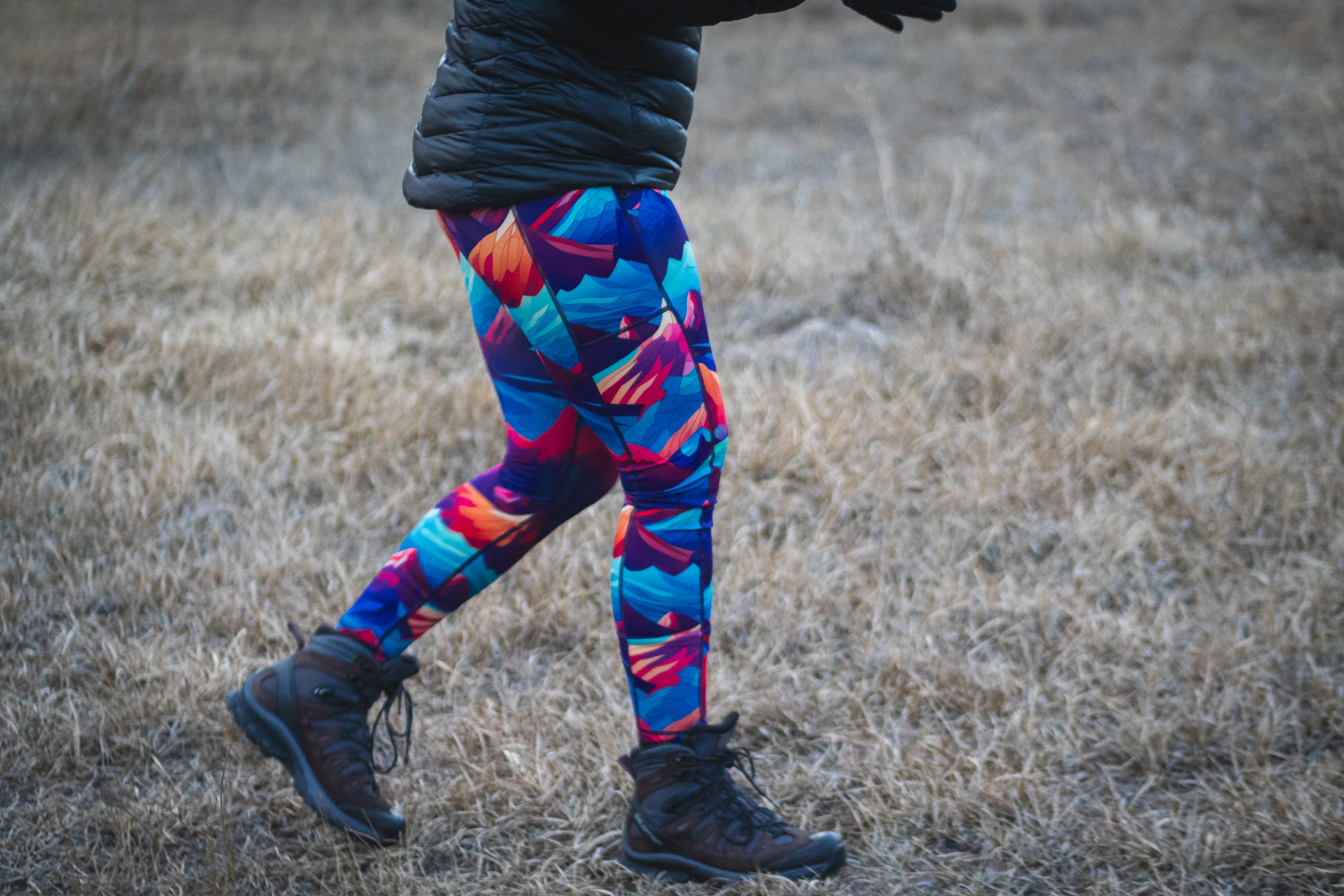 Person wearing bright colored leggings (blue, purpled, orange) that have patterns of mountains, a black puffy jacket and brown and grey high top hiking boots. The person is hiking in dried grass.