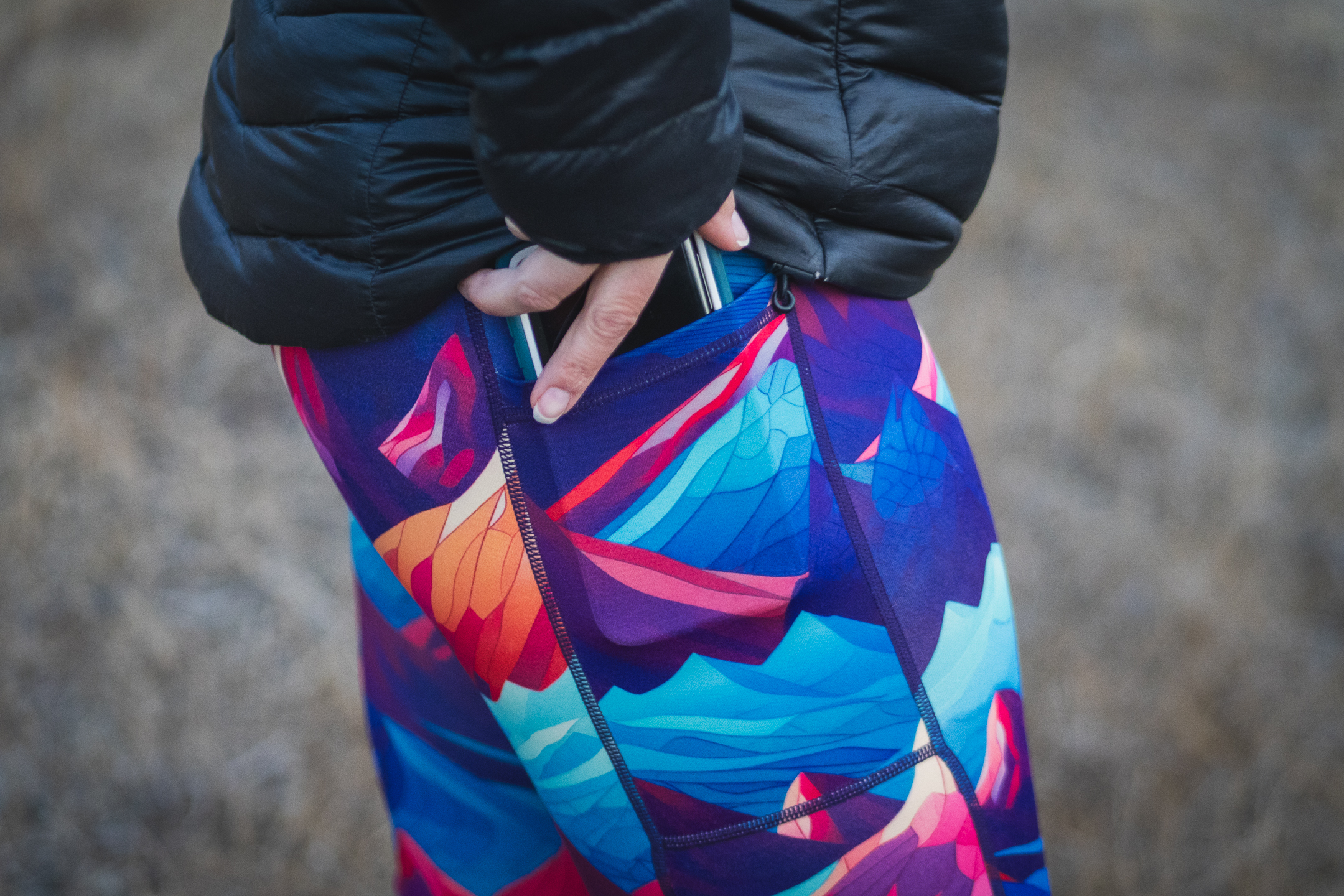 Person wearing bright colored leggings (blue, purpled, orange) that have patterns of mountains. There is a phone in the pocket, that looks to be pulled halfway out. They're either putting the phone in the pocket or taking it out.