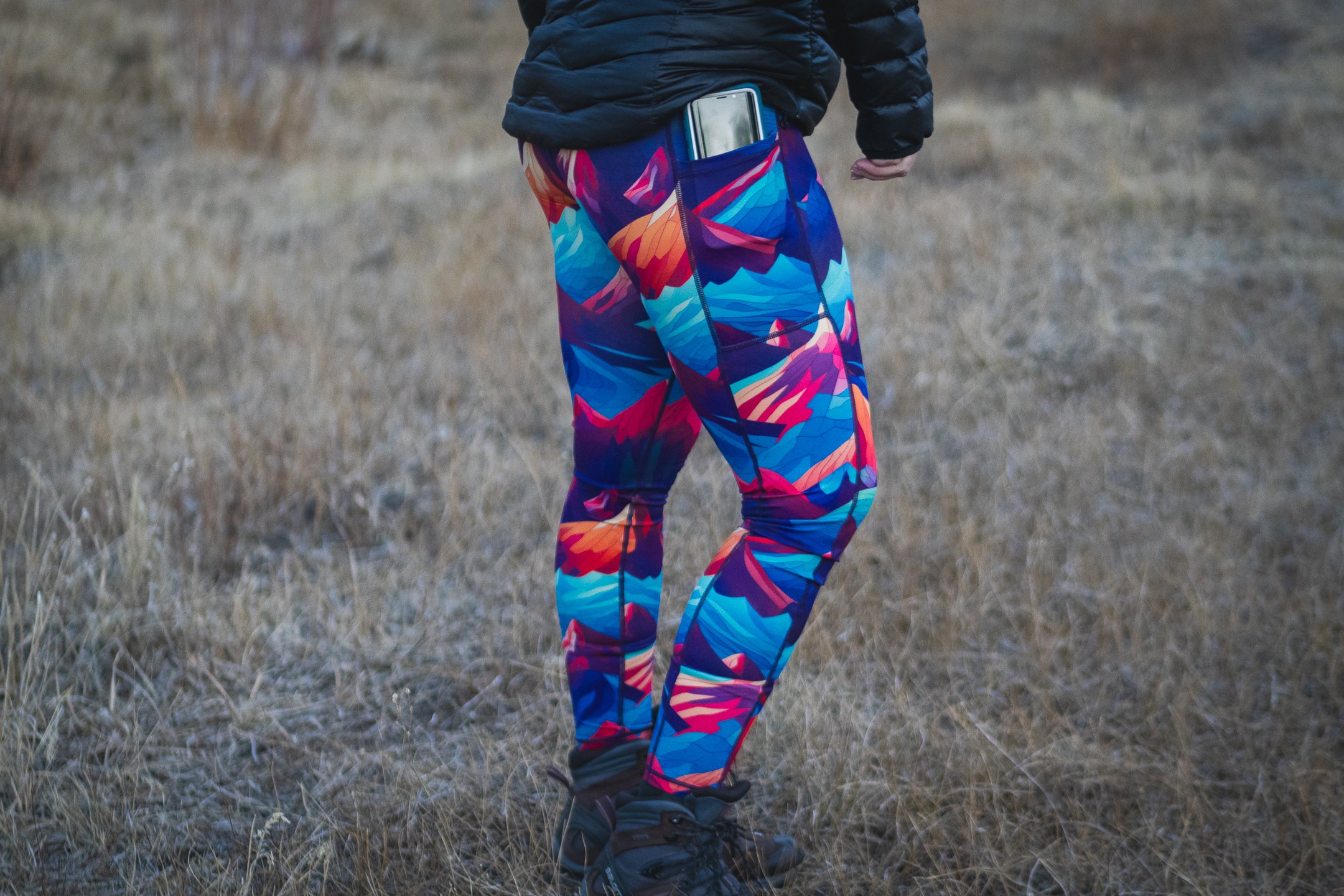 Person wearing bright colored leggings (blue, purpled, orange) that have patterns of mountains. There is a phone in the pocket, that looks to be pulled halfway out. Person is wearing hefty hiking boots and a black puffy jacket, with dried grass in the background.