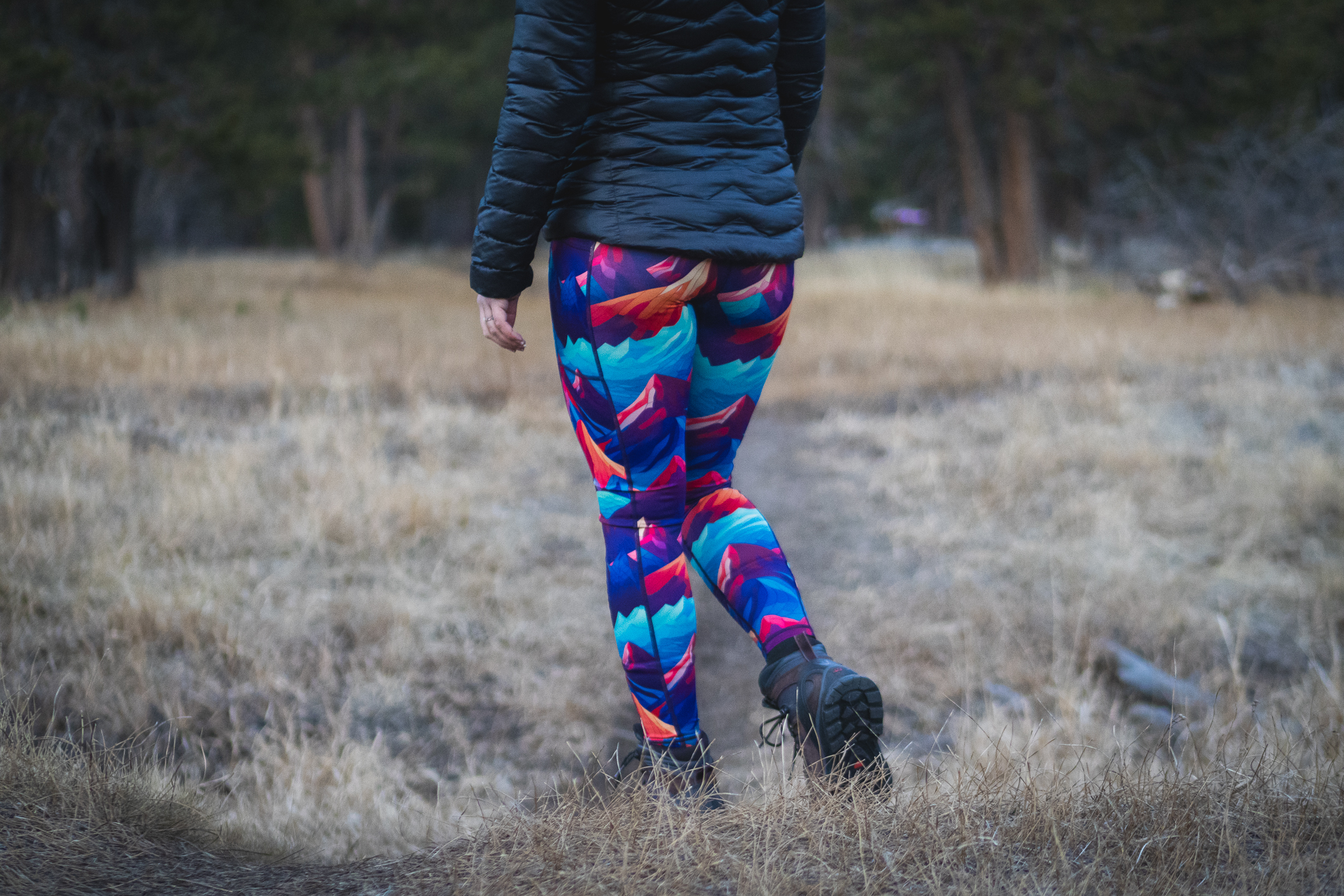 Woman in bright colored leggings (blue, purpled, orange) that have patterns of mountains, hiking on a trail in a meadow in dry grass.