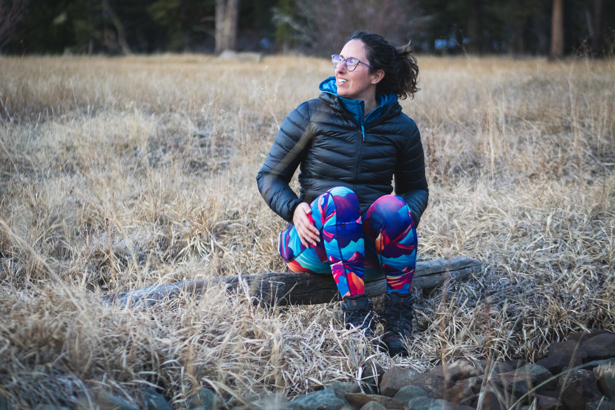 A woman sitting on a log in bright colored leggings and hiking boots looking out of the frame, smiling. He hands are resting on her legs.