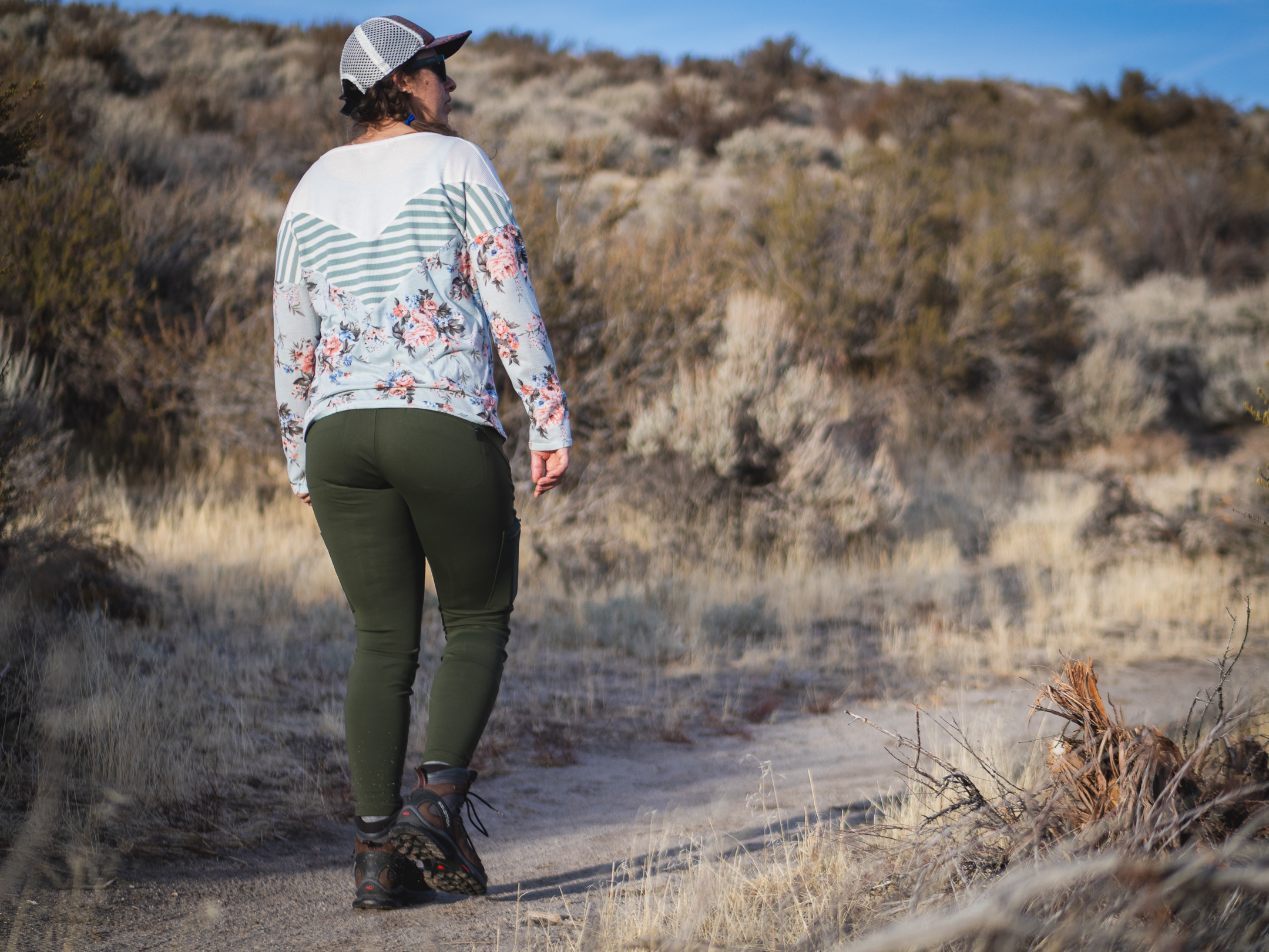 Woman hiking on dirt trail in high desert terrain, wearing forest green hiking leggings with pockets, around sunset.