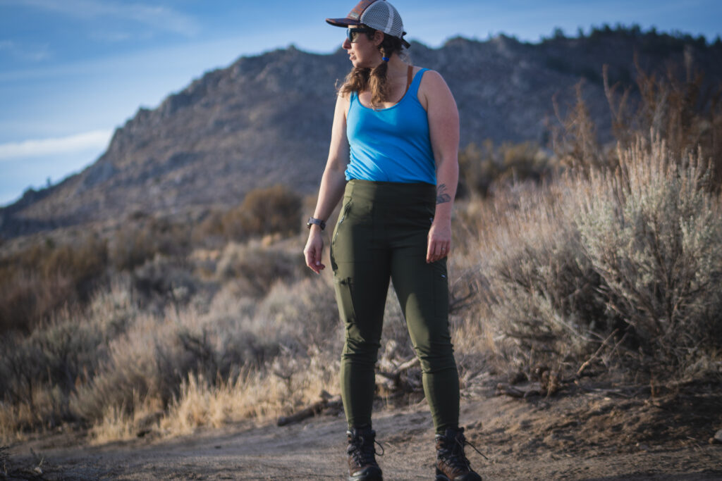 Woman hiking on dirt trail in high desert terrain, wearing forest green hiking leggings with pockets, around sunset.