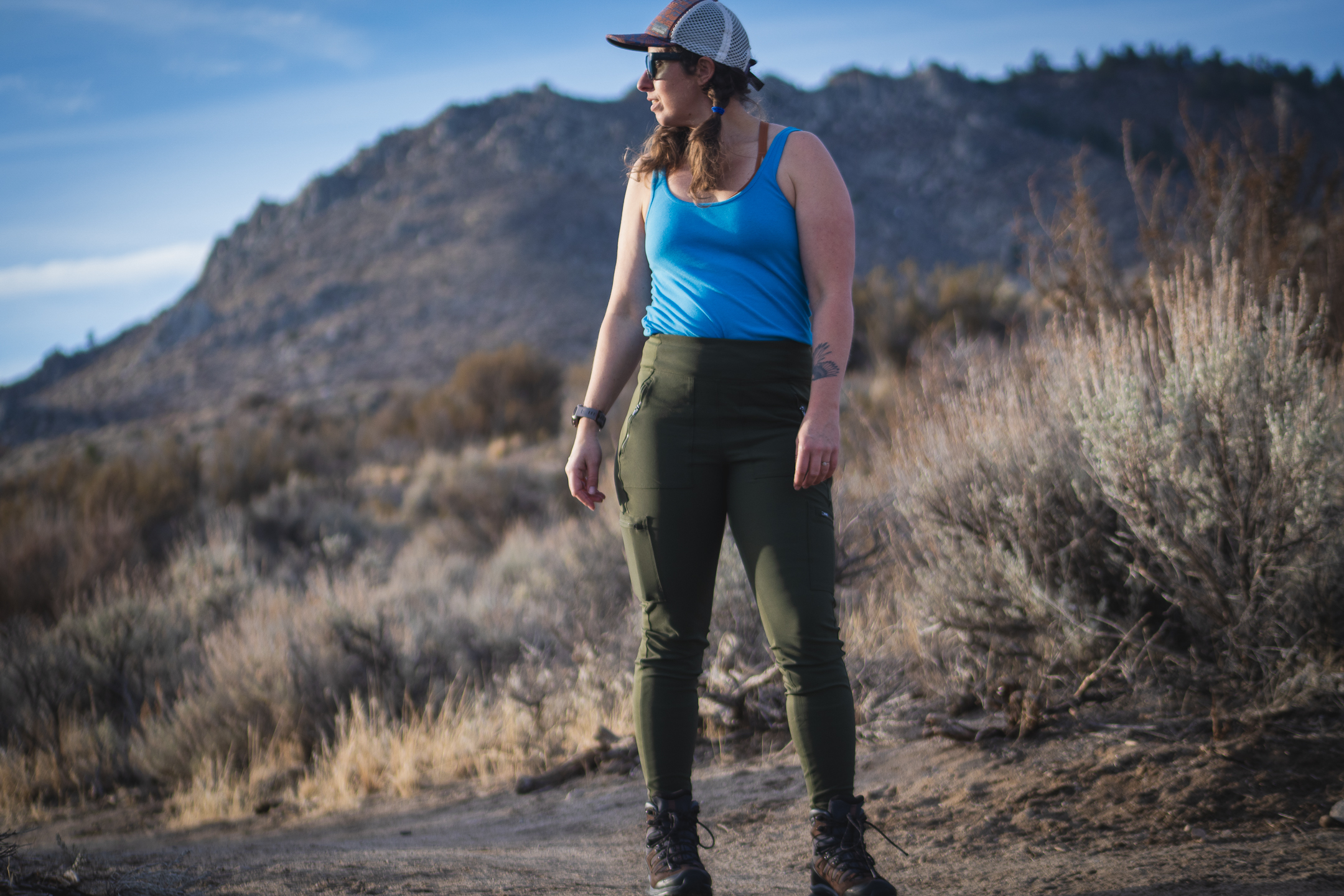 Woman hiking on dirt trail in high desert terrain, wearing forest green hiking leggings with pockets, around sunset.