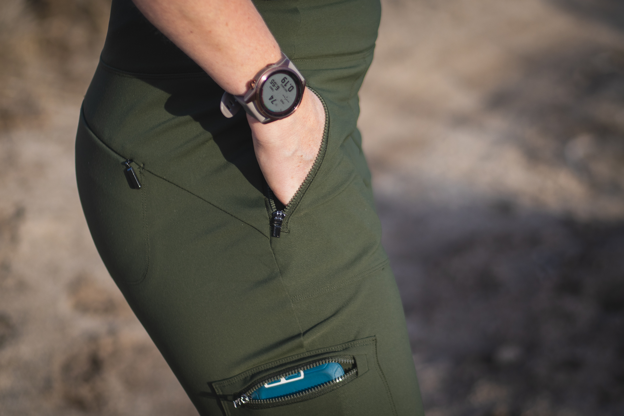 Woman hiking on dirt trail in high desert terrain, wearing forest green hiking leggings with pockets, around sunset.