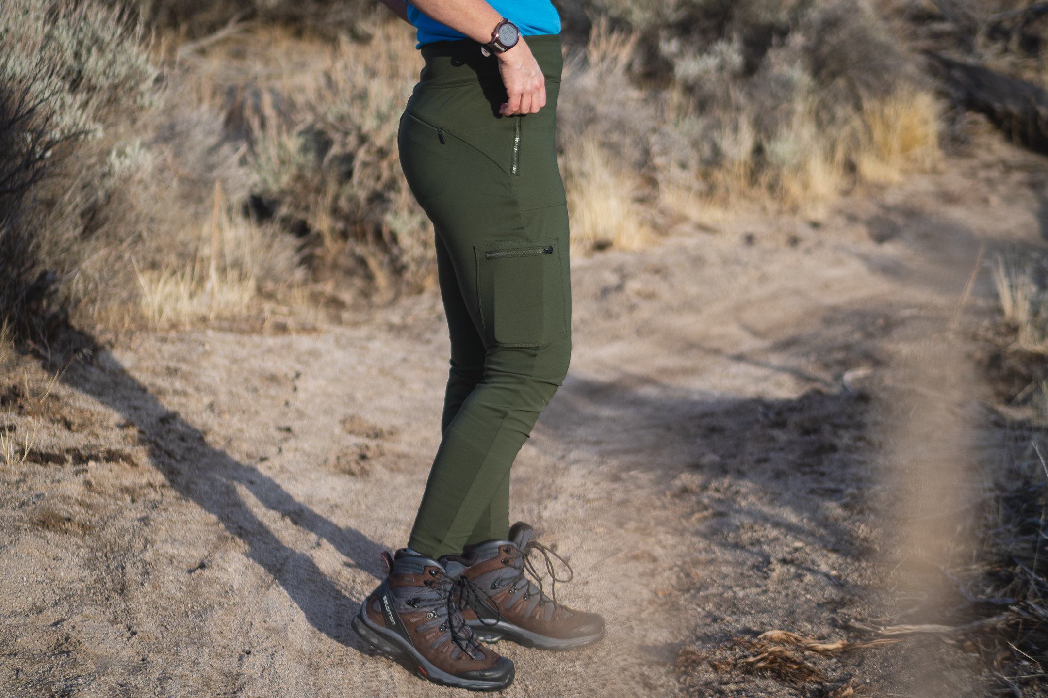 Woman hiking on dirt trail in high desert terrain, wearing forest green hiking leggings with pockets, around sunset.