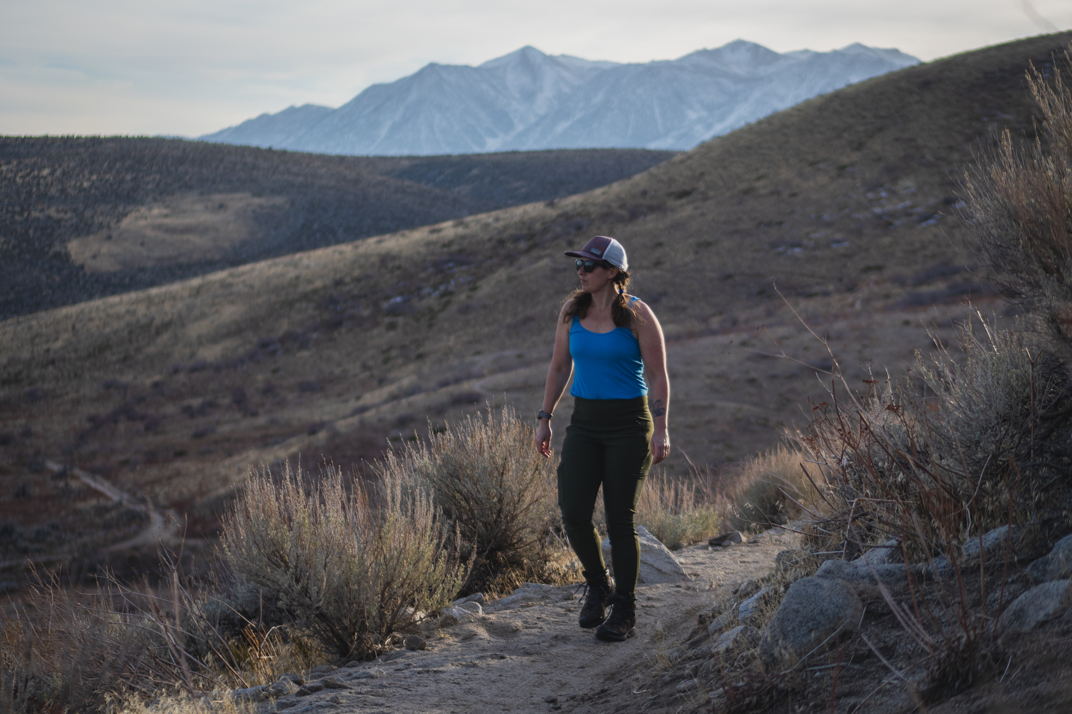 Woman hiking on dirt trail in high desert terrain, wearing forest green hiking leggings with pockets, around sunset.