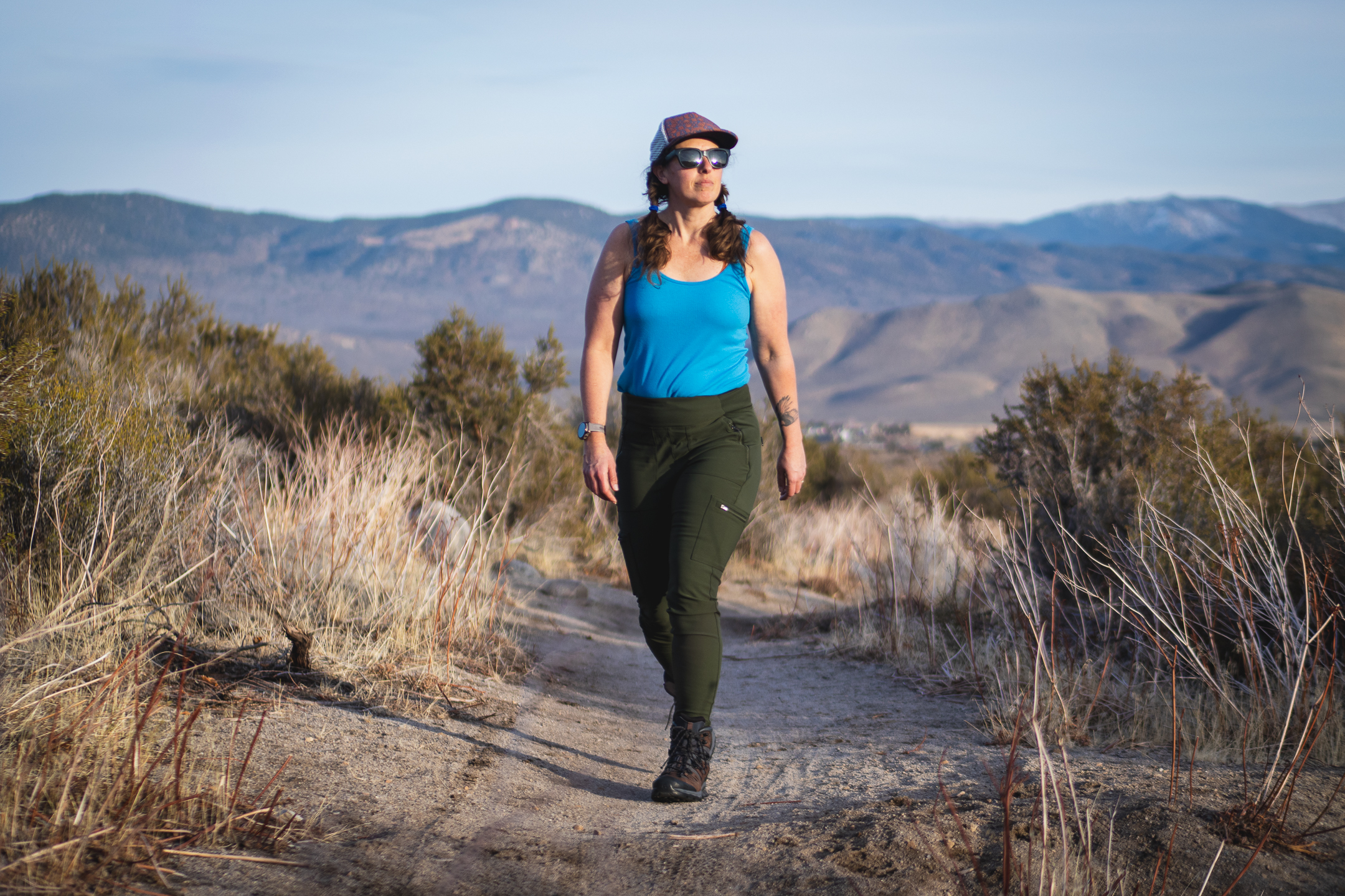 Woman hiking on dirt trail in high desert terrain, wearing forest green hiking leggings with pockets, around sunset.