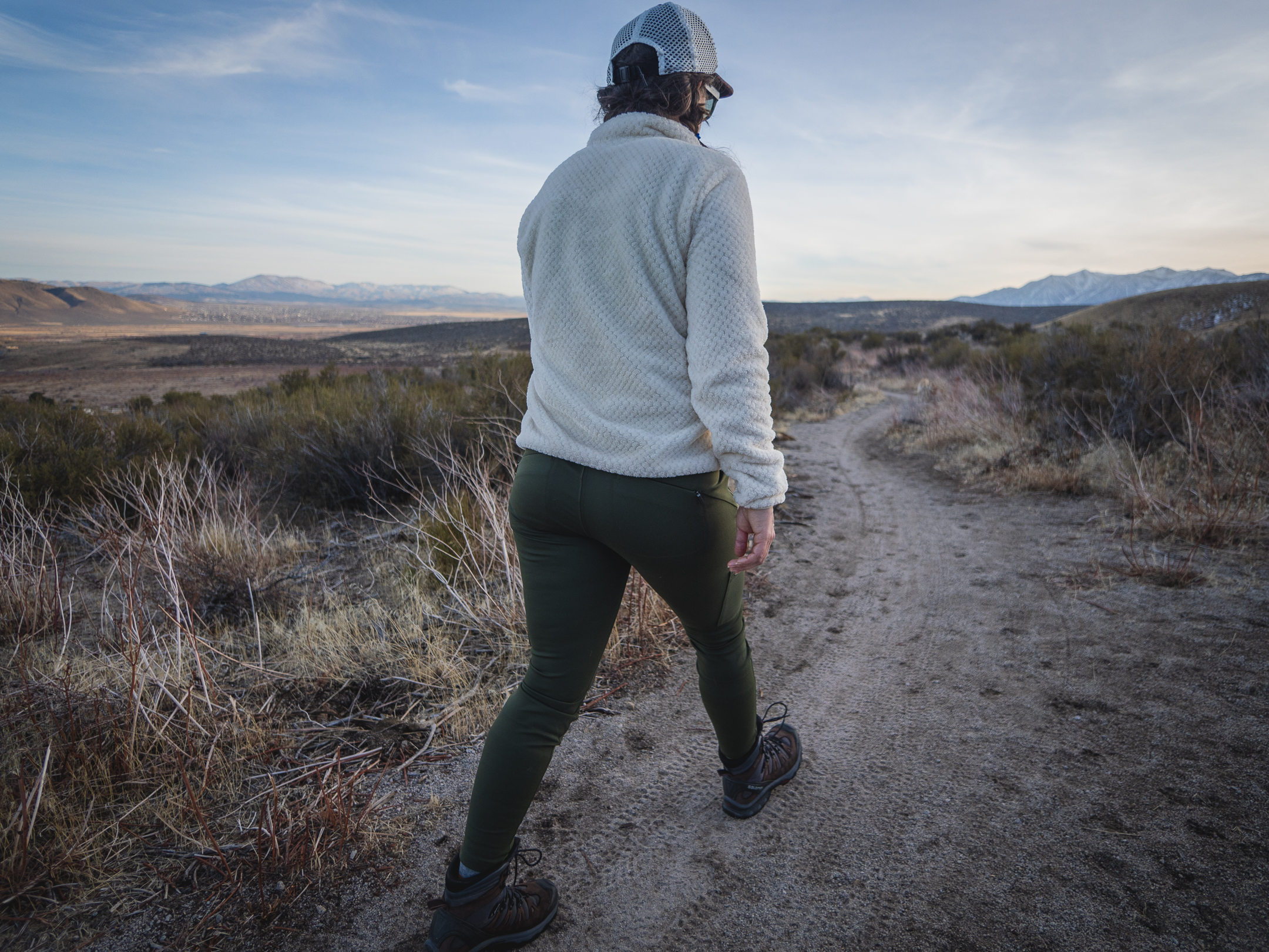 Woman hiking on dirt trail in high desert terrain, wearing forest green hiking leggings with pockets, around sunset.