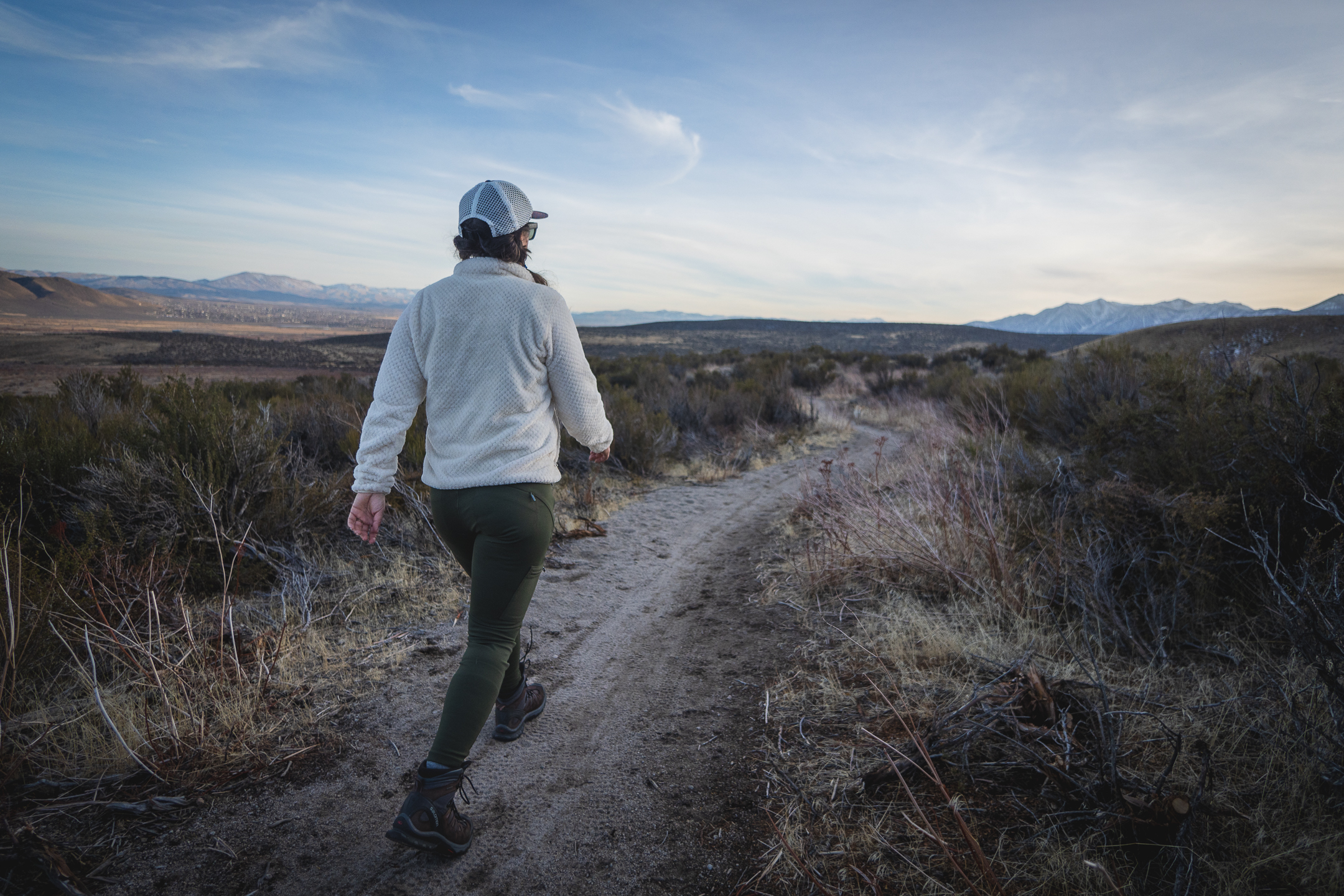 Woman hiking on dirt trail in high desert terrain, wearing forest green hiking leggings with pockets, around sunset.