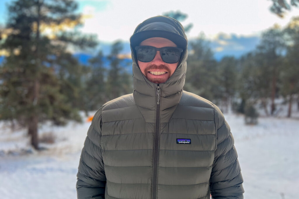 Front view of a person smiling while wearing the Patagonia Down Sweater Hoody with the hood fully zipped, standing in a snowy landscape with trees in the background.