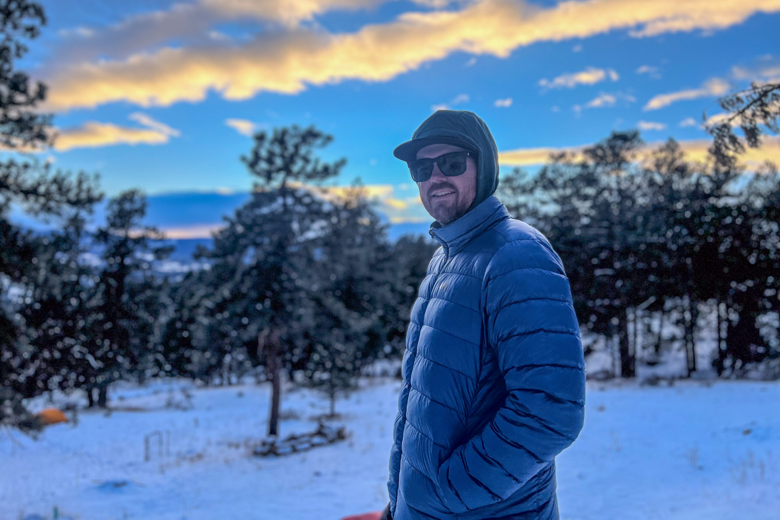 A man in an REI 650 Down Jacket, wearing sunglasses and a cap, looking into the distance with snowy trees and a vivid sunset behind him.