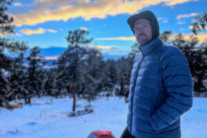 A man in a blue REI 650 Down Jacket stands with his hands in his pockets, gazing off into the snowy forest during an early winter evening.