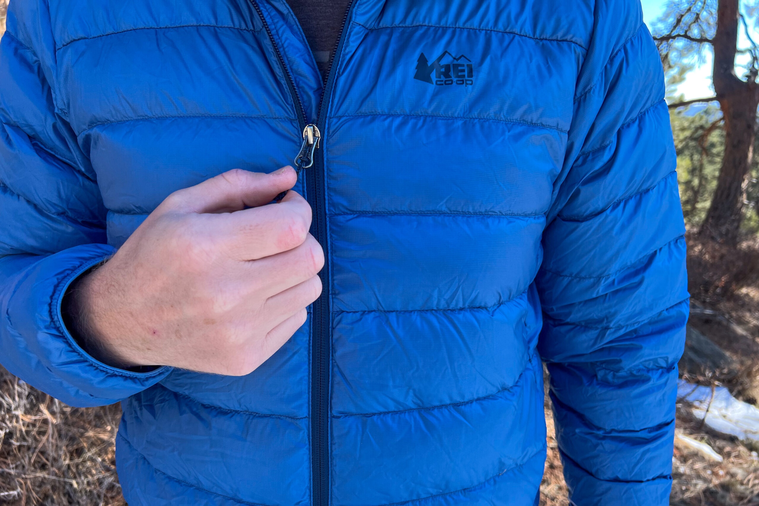 Close-up of a person’s hand holding the front zipper of the REI 650 Down Jacket, focusing on the fabric texture and stitching details.