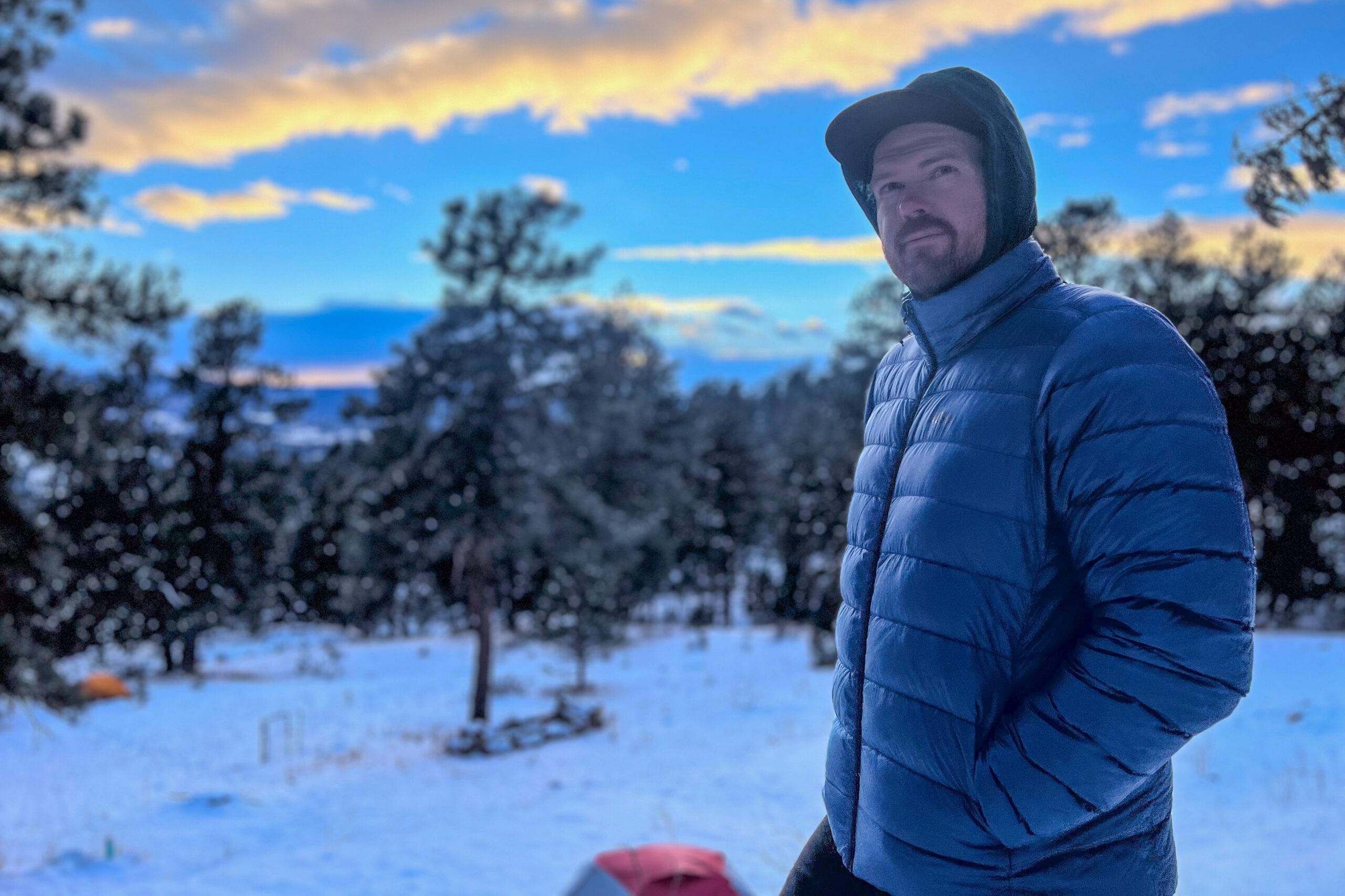 A man in a blue REI 650 Down Jacket stands with his hands in his pockets, gazing off into the snowy forest during an early winter evening.