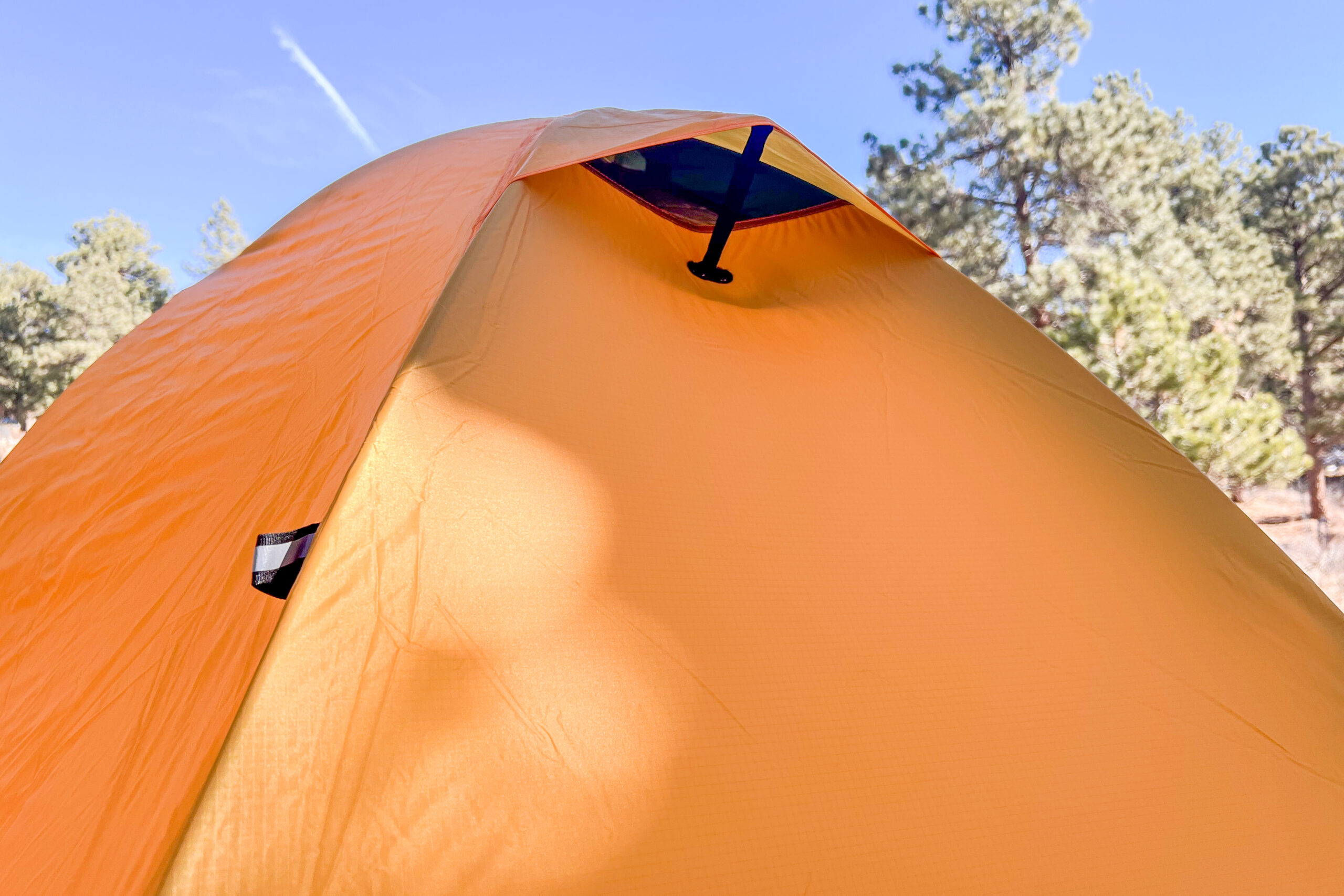 An orange tent in a forested setting. The tent has a kickstand style vent in the fly.