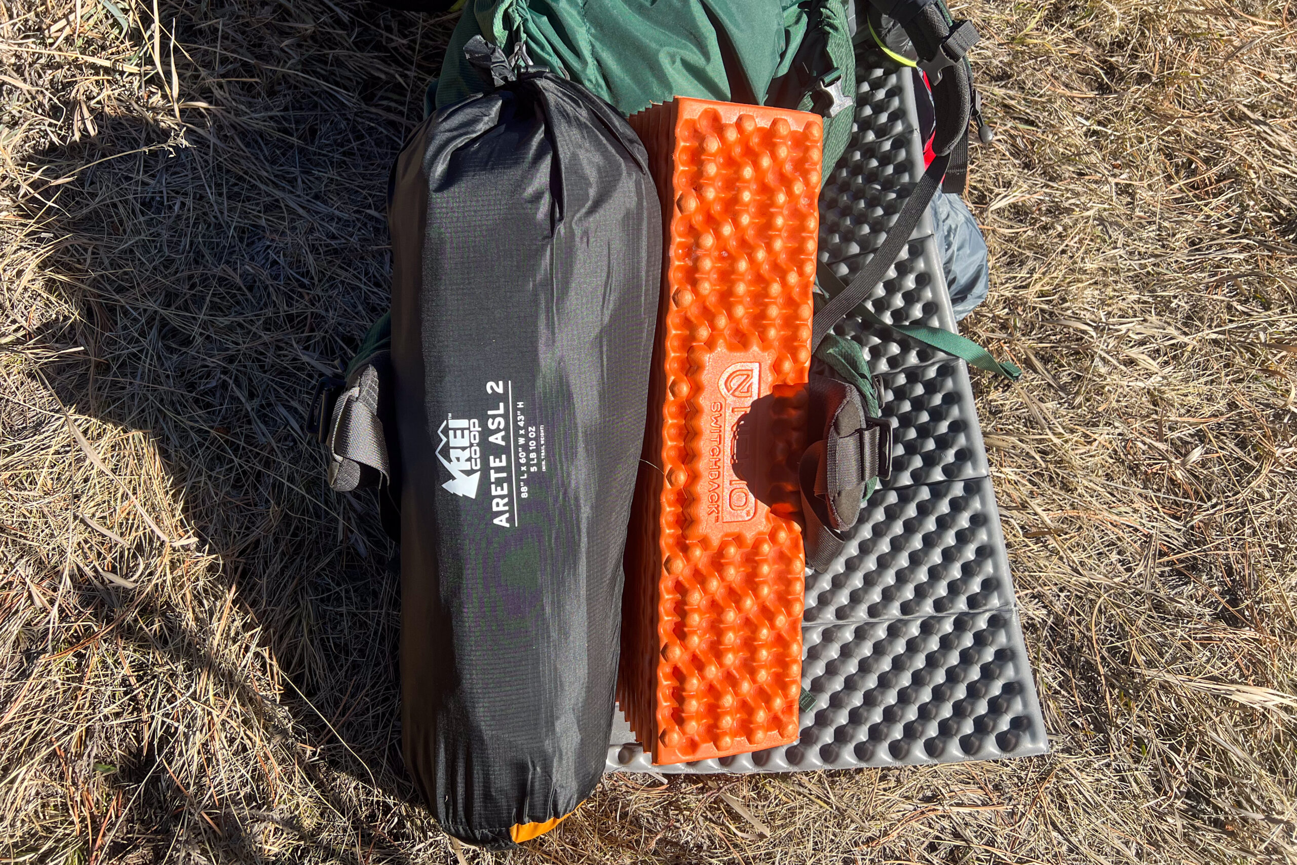 A tent packed into it's stuff sack and laying on the ground next to a foam sleeping mat.