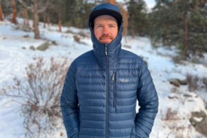 A person standing in snowy terrain wearing the Rab Microlight Alpine jacket fully zipped with the hood up for maximum protection.