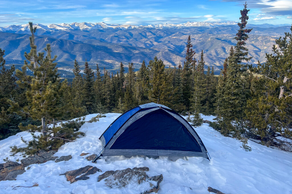 The Samaya 2.0 tent pitched on snow with evergreen trees and panoramic views of rugged mountain peaks in the background.