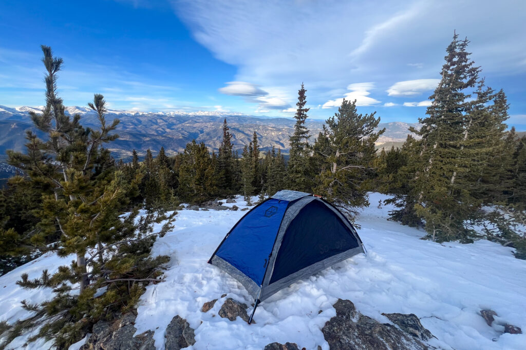 A blue and silver Samaya 2.0 tent pitched on snow with a stunning backdrop of mountain ranges under a blue sky.