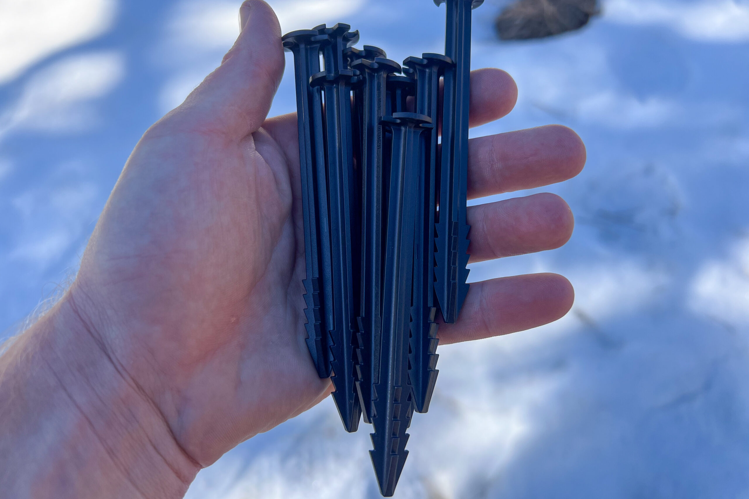 A hand holding several black Samaya 2.0 stakes with serrated edges, resting against a snowy backdrop.