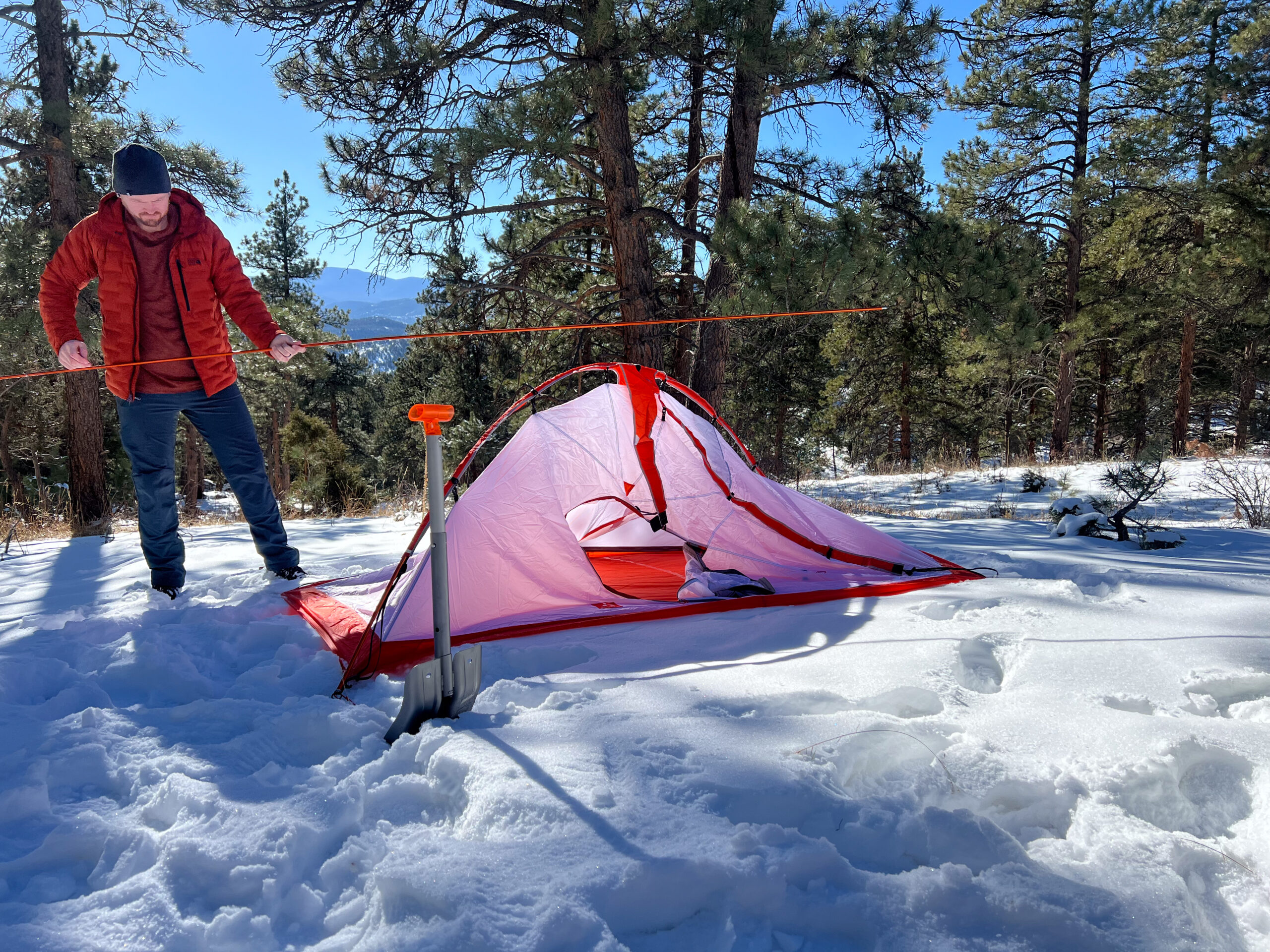 A man in an orange coat pitching a tent in the snow surrounded by pine trees.