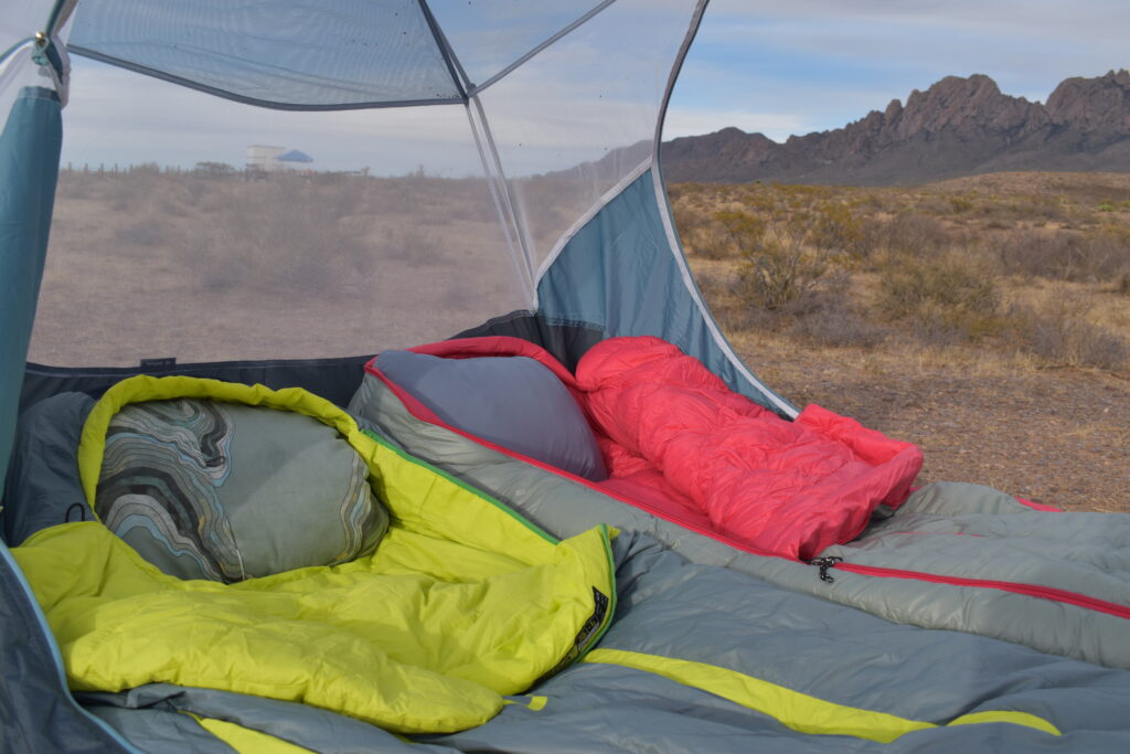 An open tent shows the Therm-a-rest Compressible Pillow and HEST Camp Pillows on sleeping bags with rugged mountains in the background