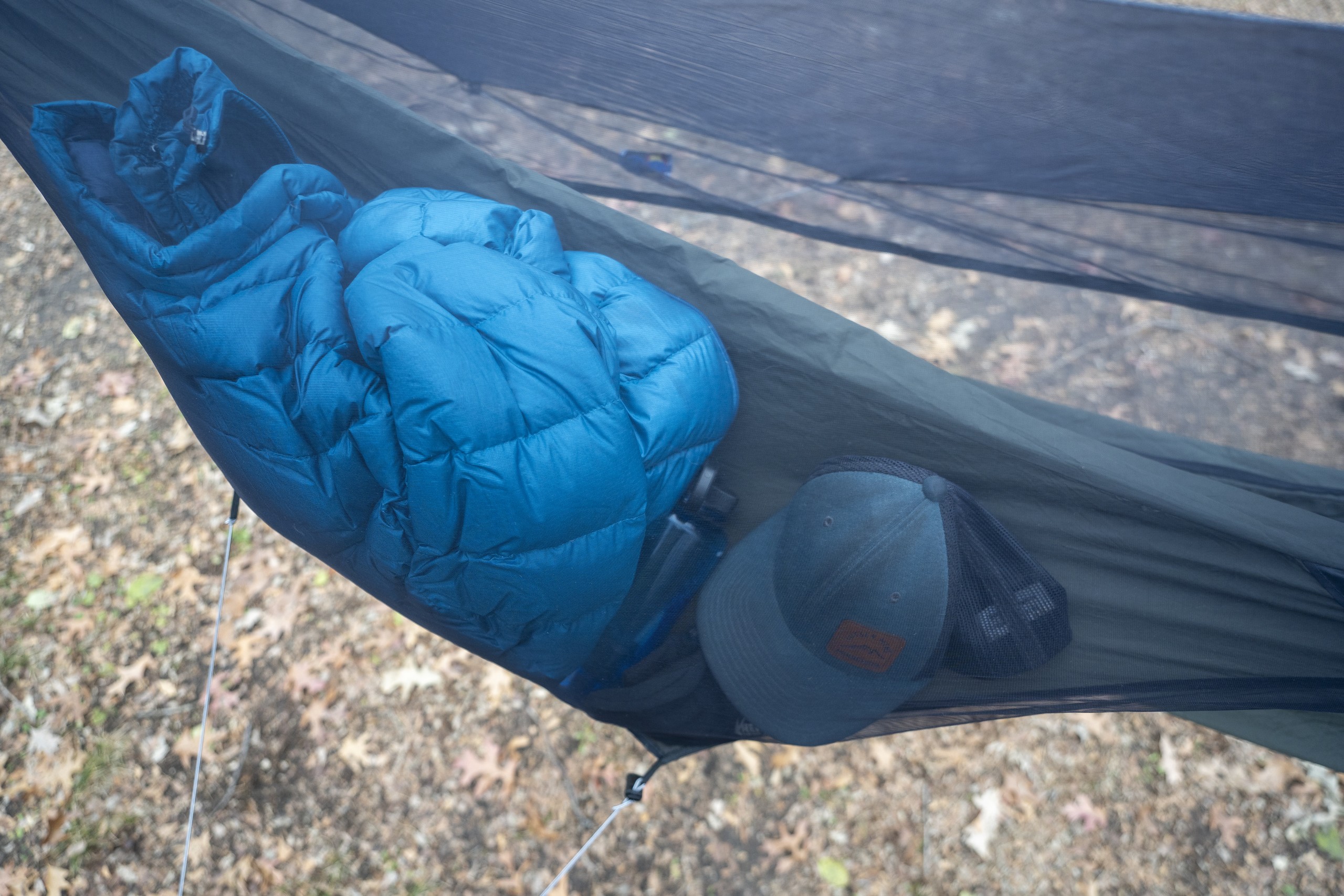 A side pocket on a hammock holds a blue jacket, baseball hat, and other small items