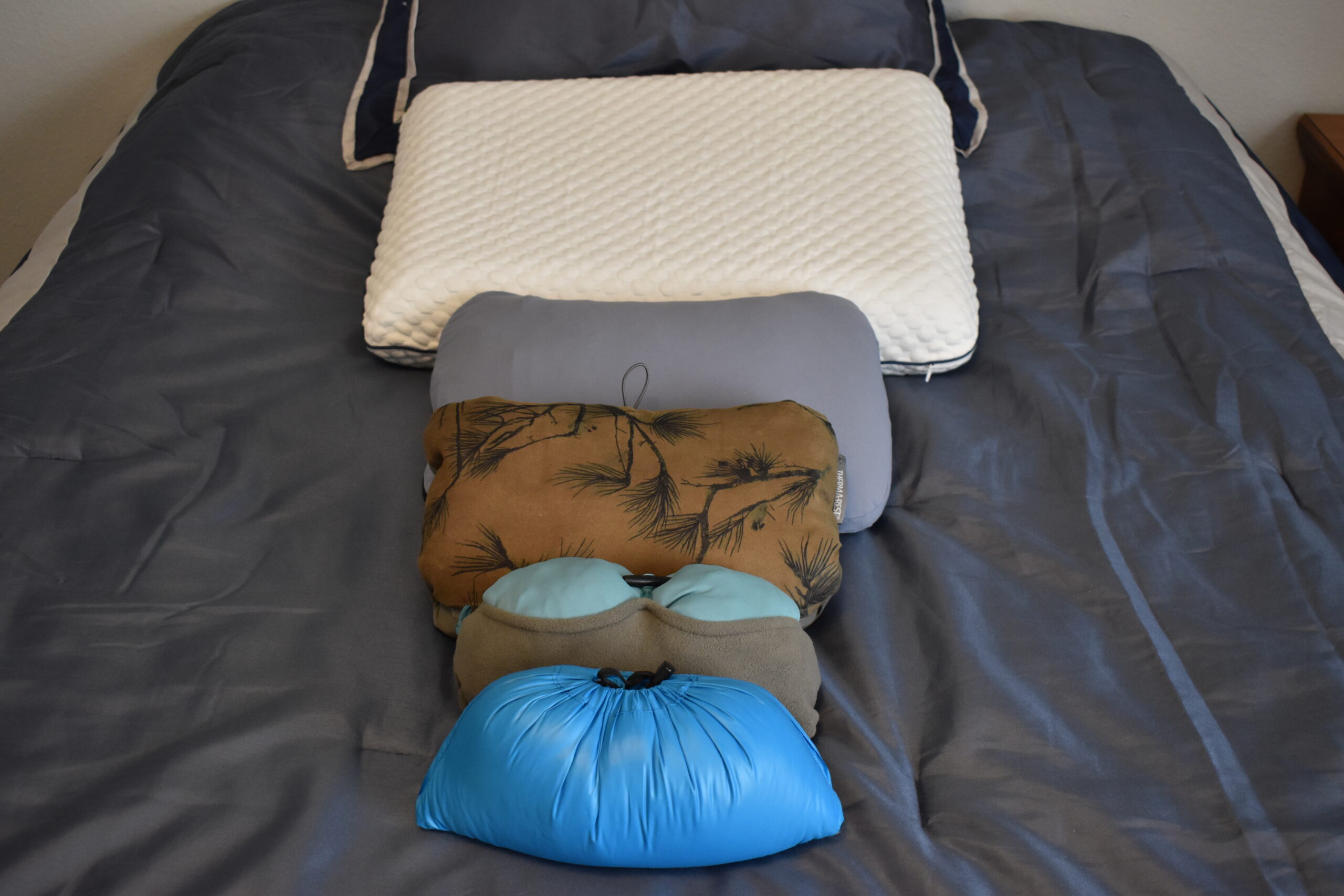 A line of pillows on a bed shows the foam pillows we tested from the largest to smallest: Weekender Gel Memory Foam Pillow > HEST Camp Pillow > Therm-a-rest Compressible Pillow > REI Trailmade Mummy Pillow > Western Mountaineering Cloudrest Pillow