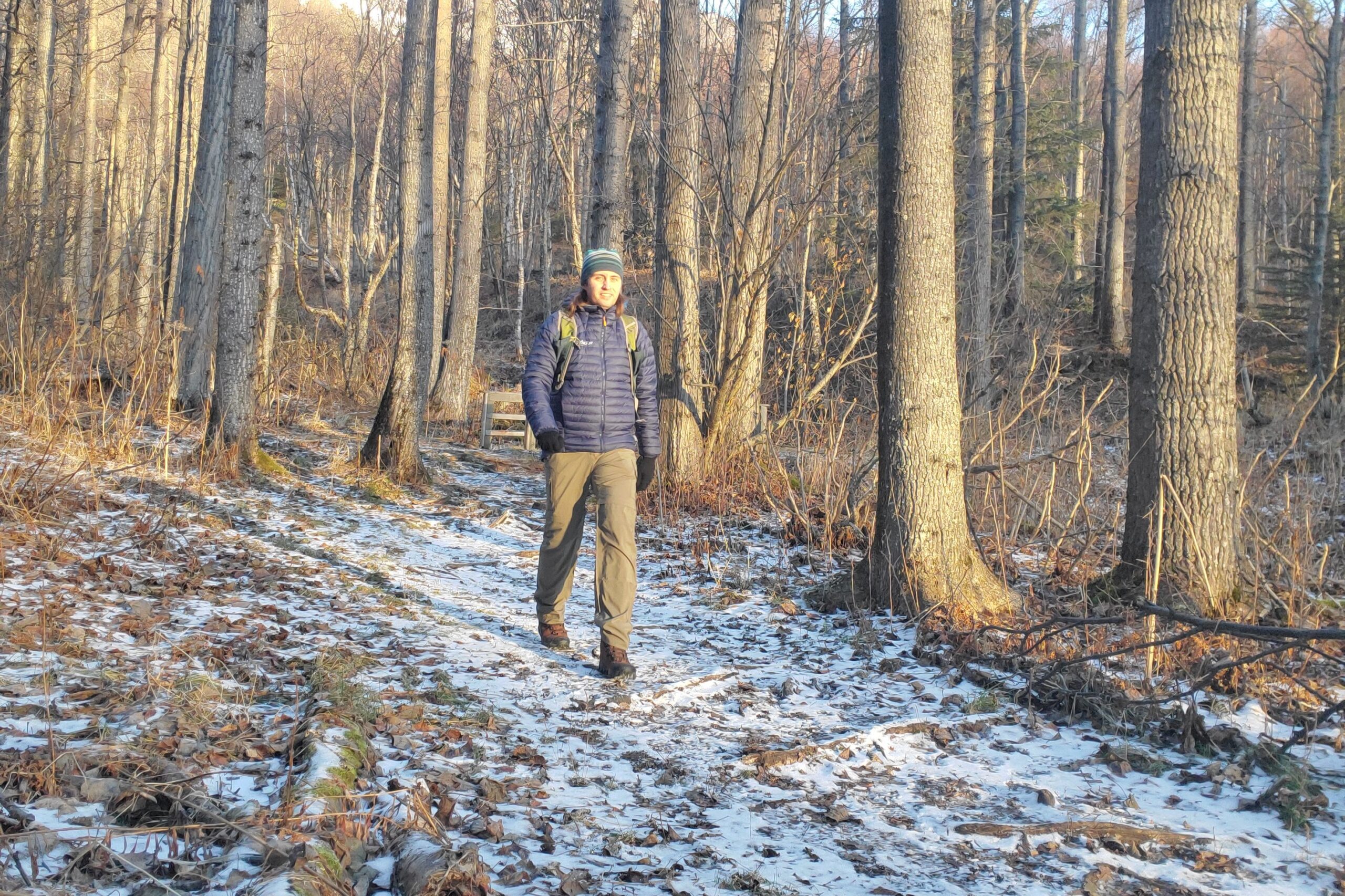 A man hikes down a lightly snowy trail in a blue jacket and green pants.