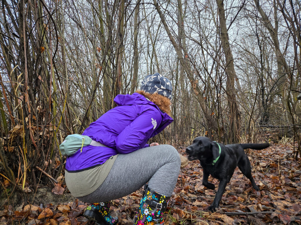 a woman croutched down saying hi to her black lab dog in the forest while wearing a fanny pack