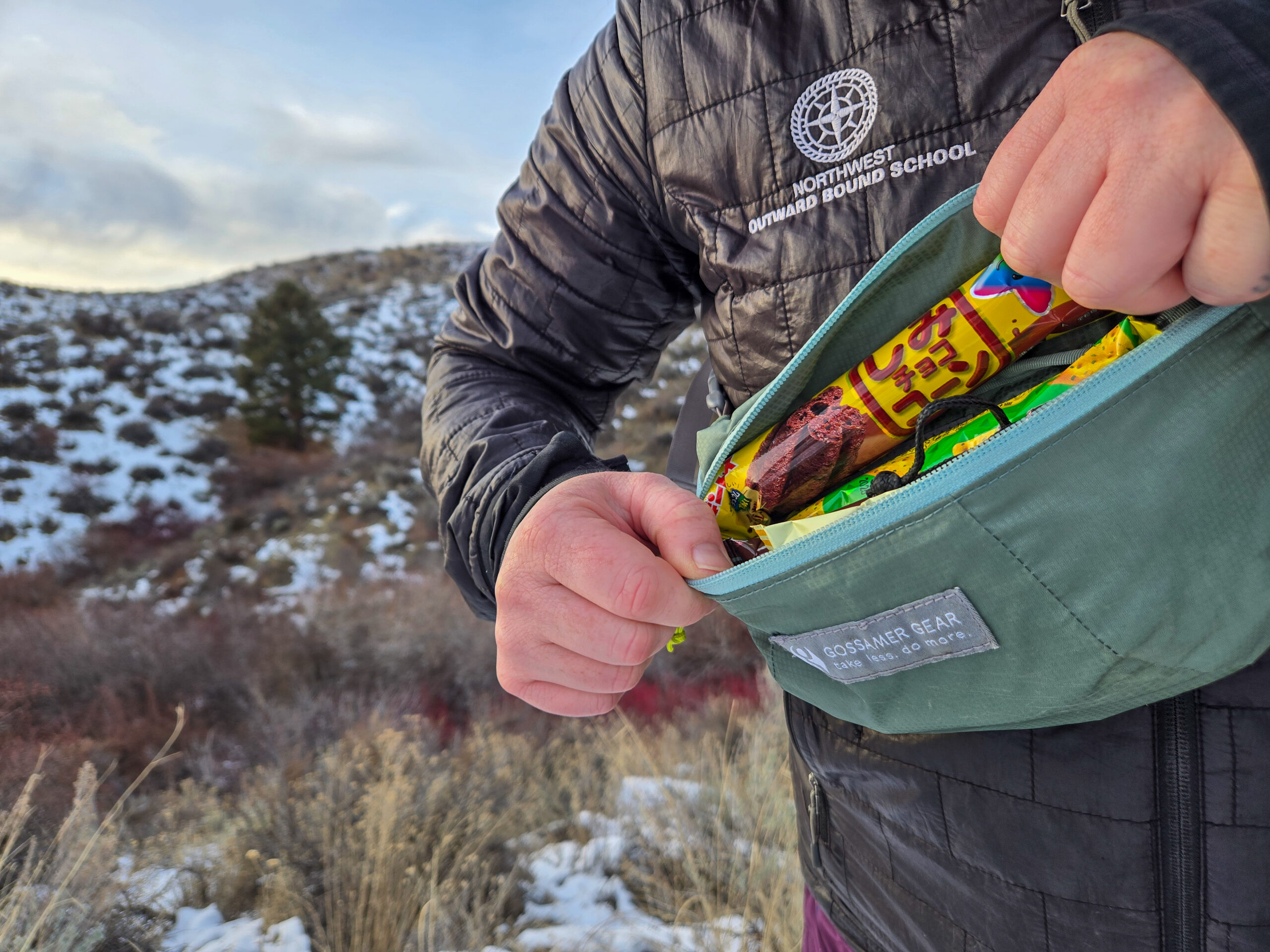 a person unzipping the bumster to grab a snack from inside the fanny pack on a snowy hike