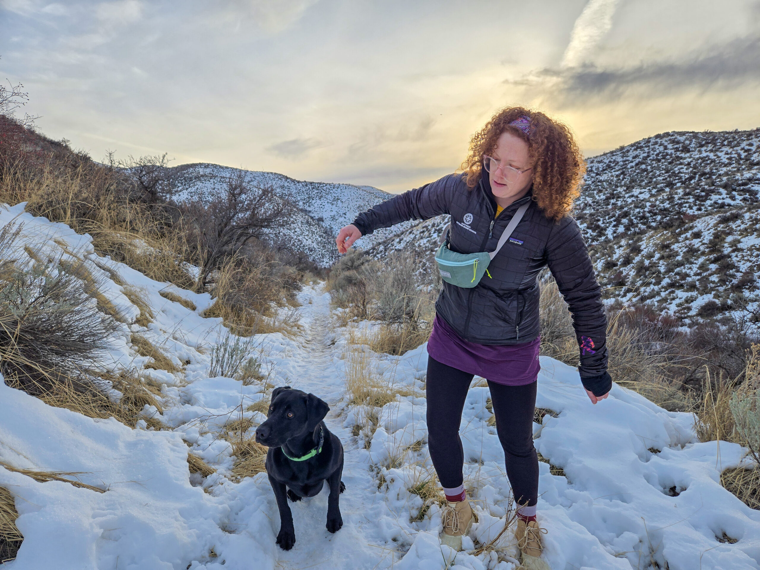 a woman walking on a a snowy hillside with her dog with the sun setting while wearing a fanny pack by gossamer.