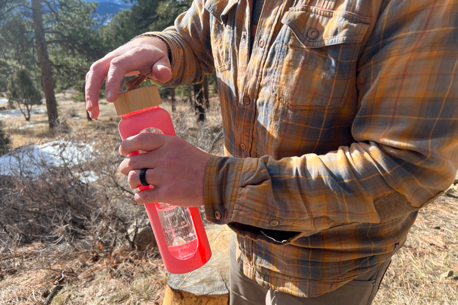 The image shows a person about to open a water bottle in a forested setting.