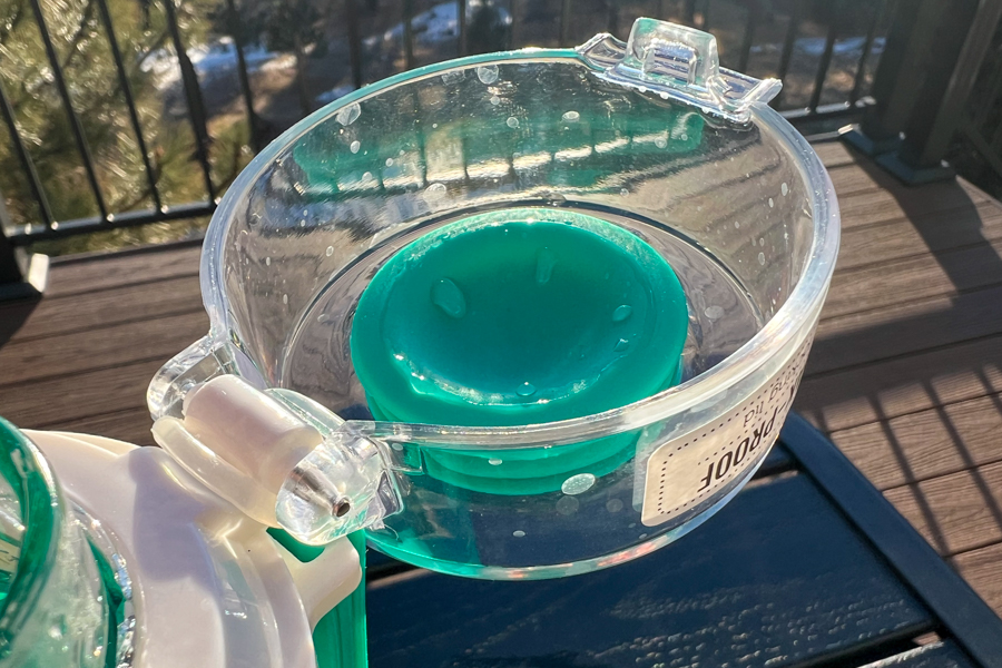 Close-up of a green silicone funnel inside the clear lid of a glass water bottle, set on a wooden table outdoors.